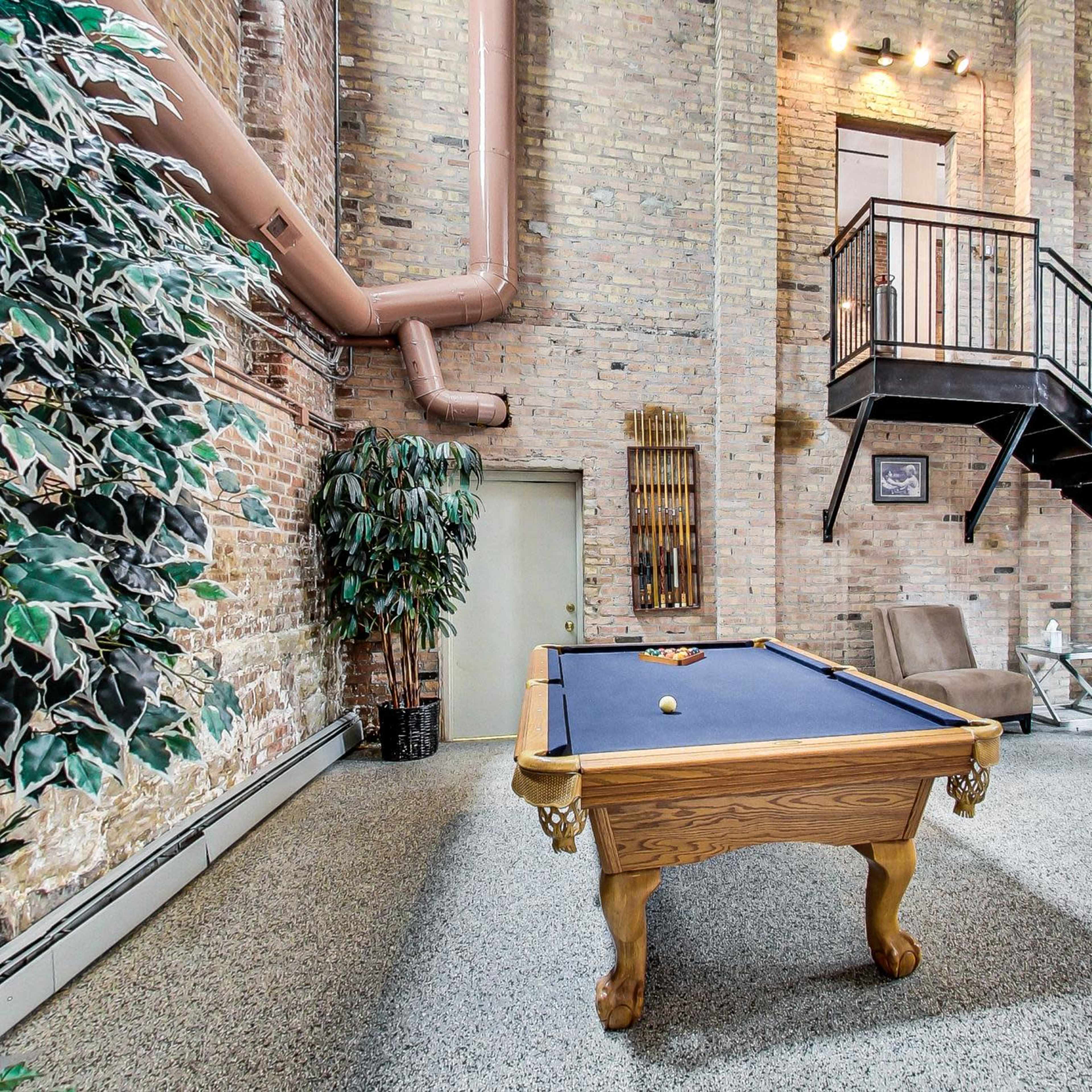 A spacious room with exposed brick walls, featuring a pool table in the center and a staircase leading to a second level.