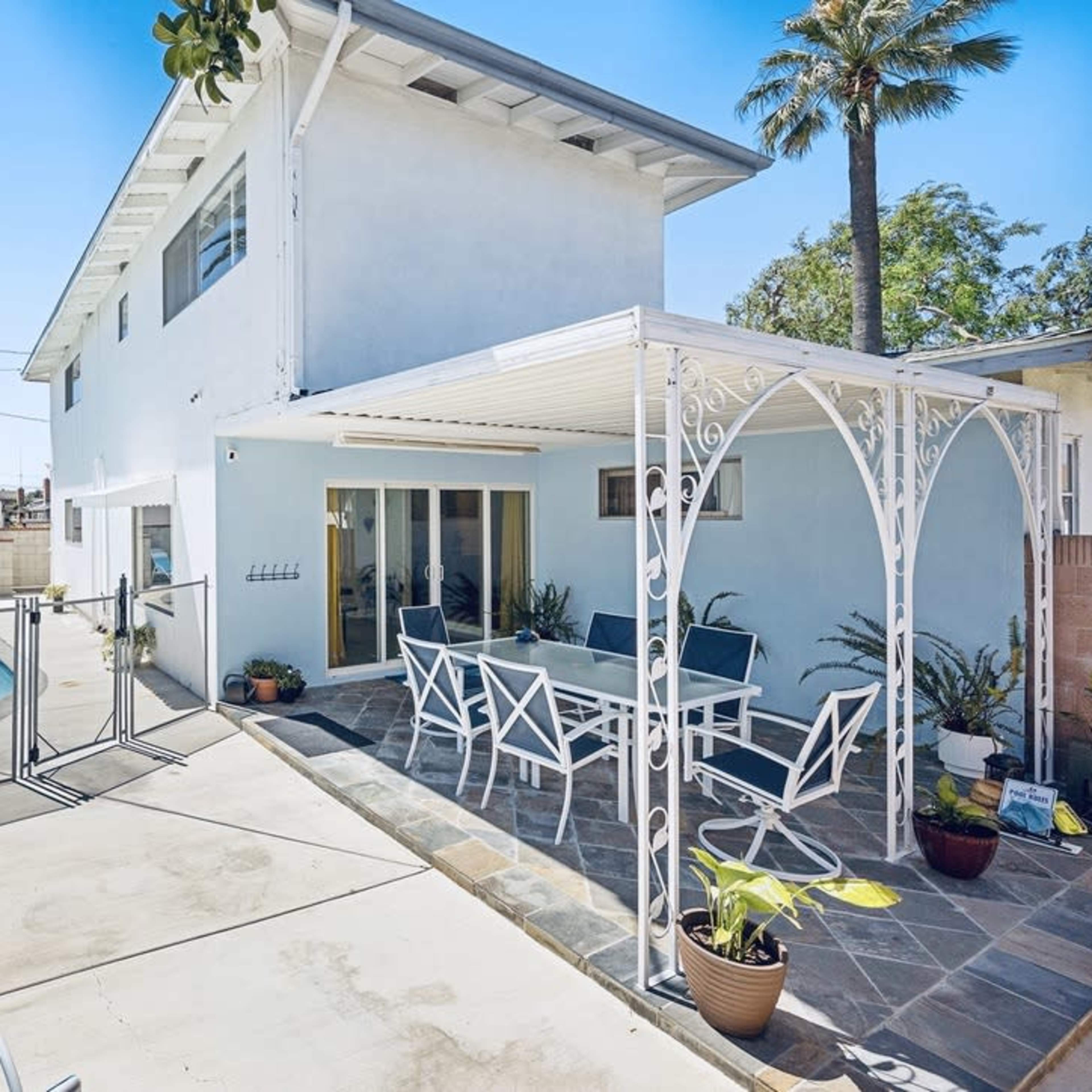 A backyard features a swimming pool, a patio with a white metal table and chairs, and palm trees surrounding a two-story blue house.