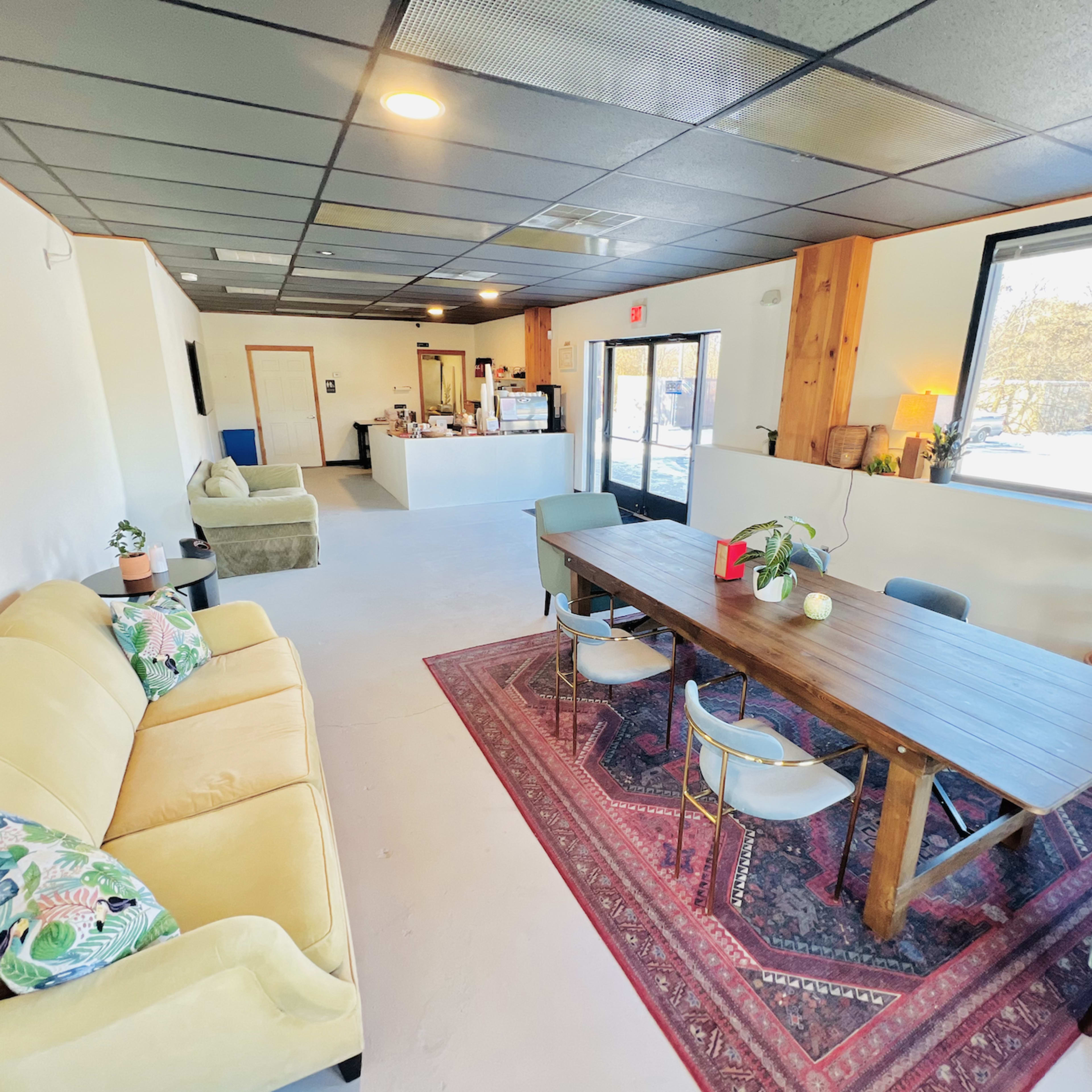 A spacious, brightly lit common area with a large wooden table surrounded by chairs, a sofa, and a kitchenette in the background.