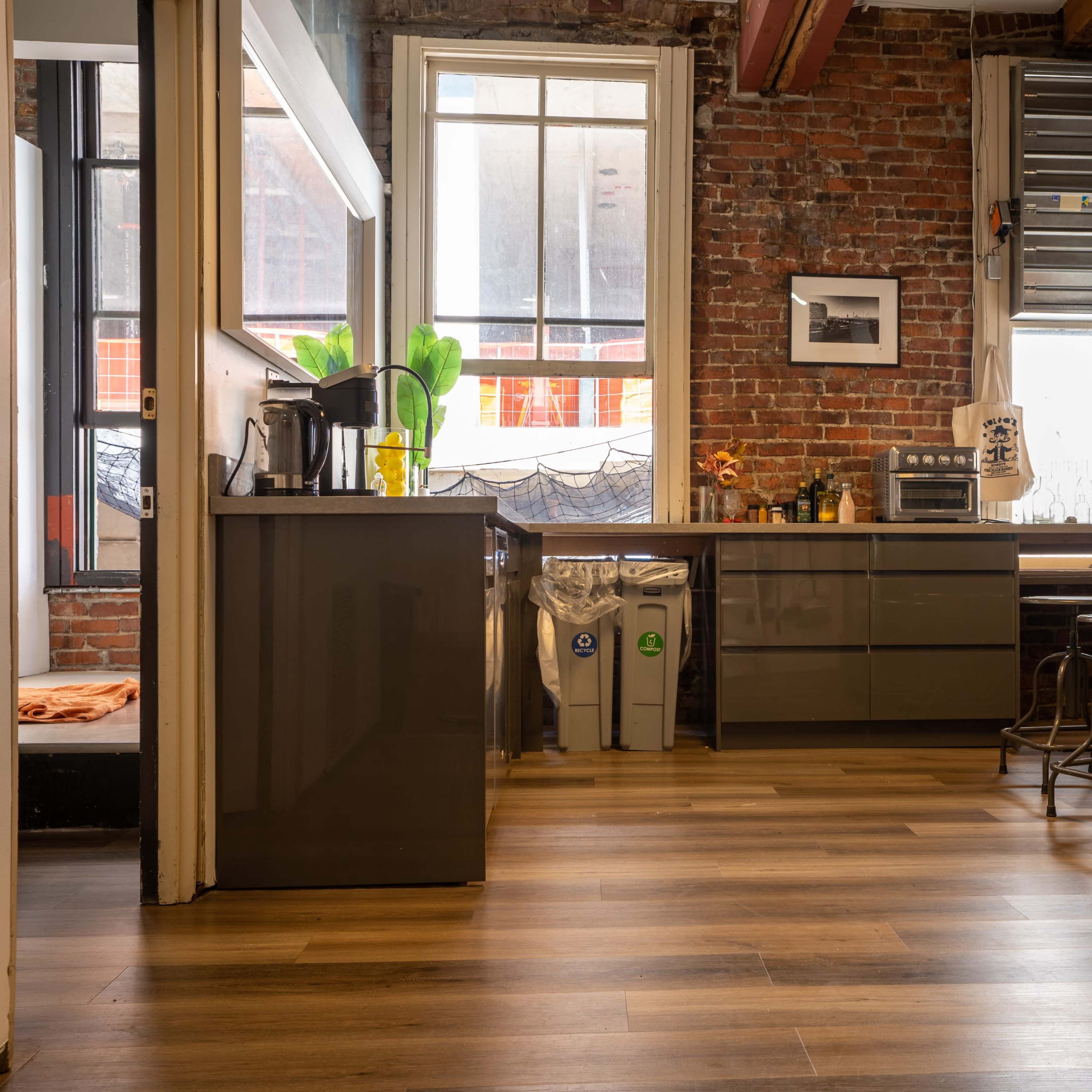 A modern kitchen with a gray countertop, stainless steel refrigerator, and large windows revealing exposed brick walls.