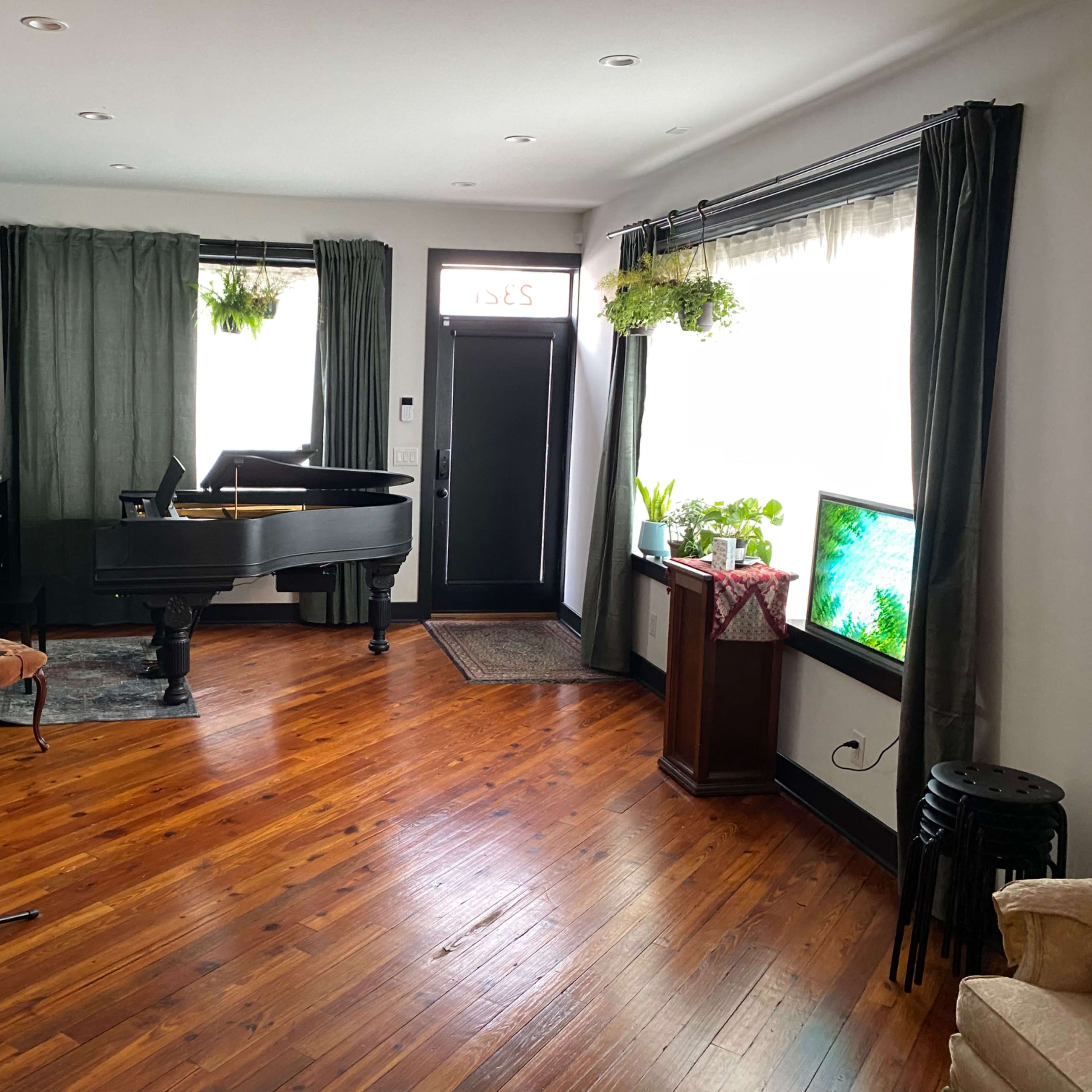 A living room featuring a grand piano, a microphone stand, potted plants, and a television, with hardwood flooring and large windows.