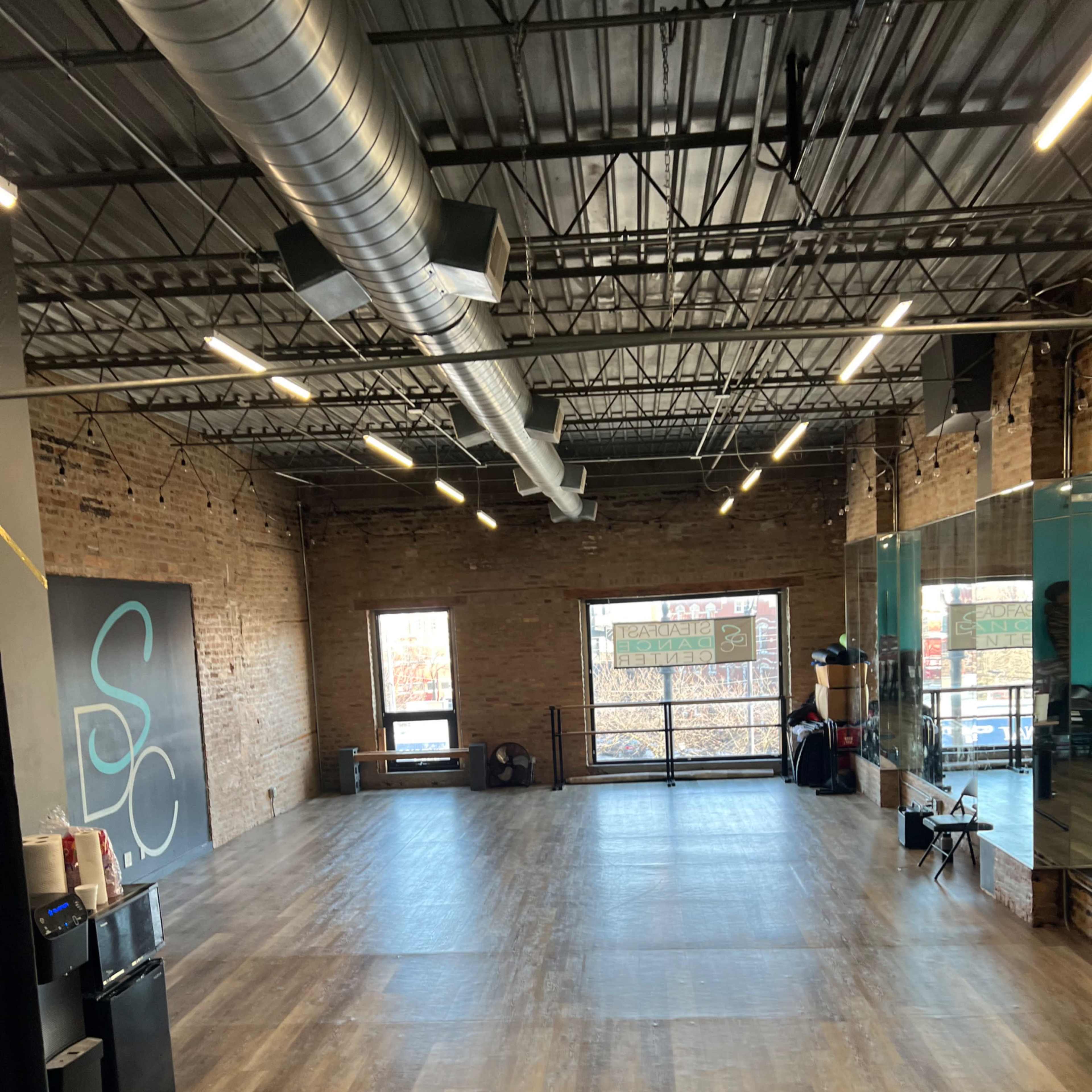A spacious, empty room with wood flooring, exposed brick walls, and large windows along one side.