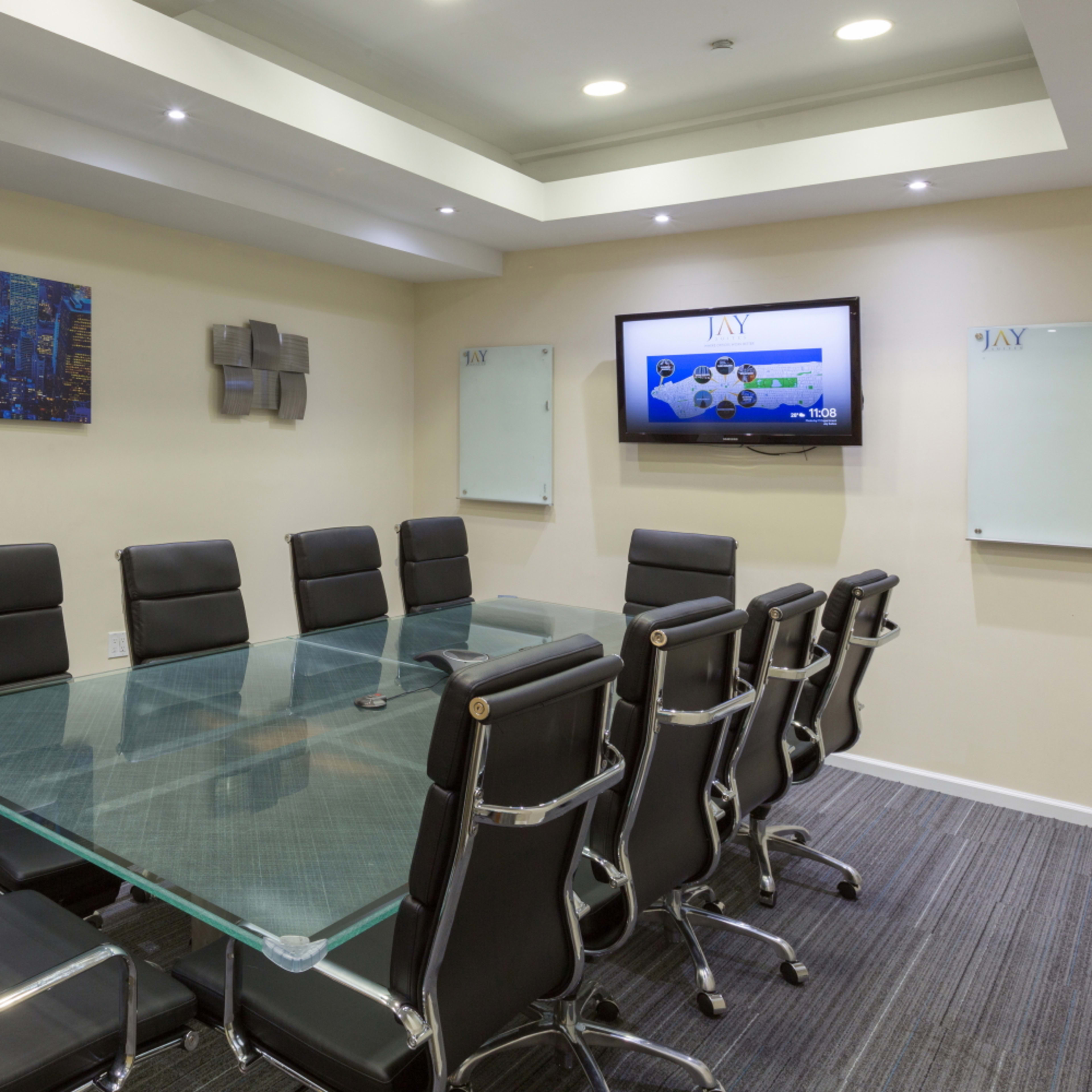 A modern conference room features a large glass table surrounded by black ergonomic chairs, with digital displays and whiteboards on the walls.