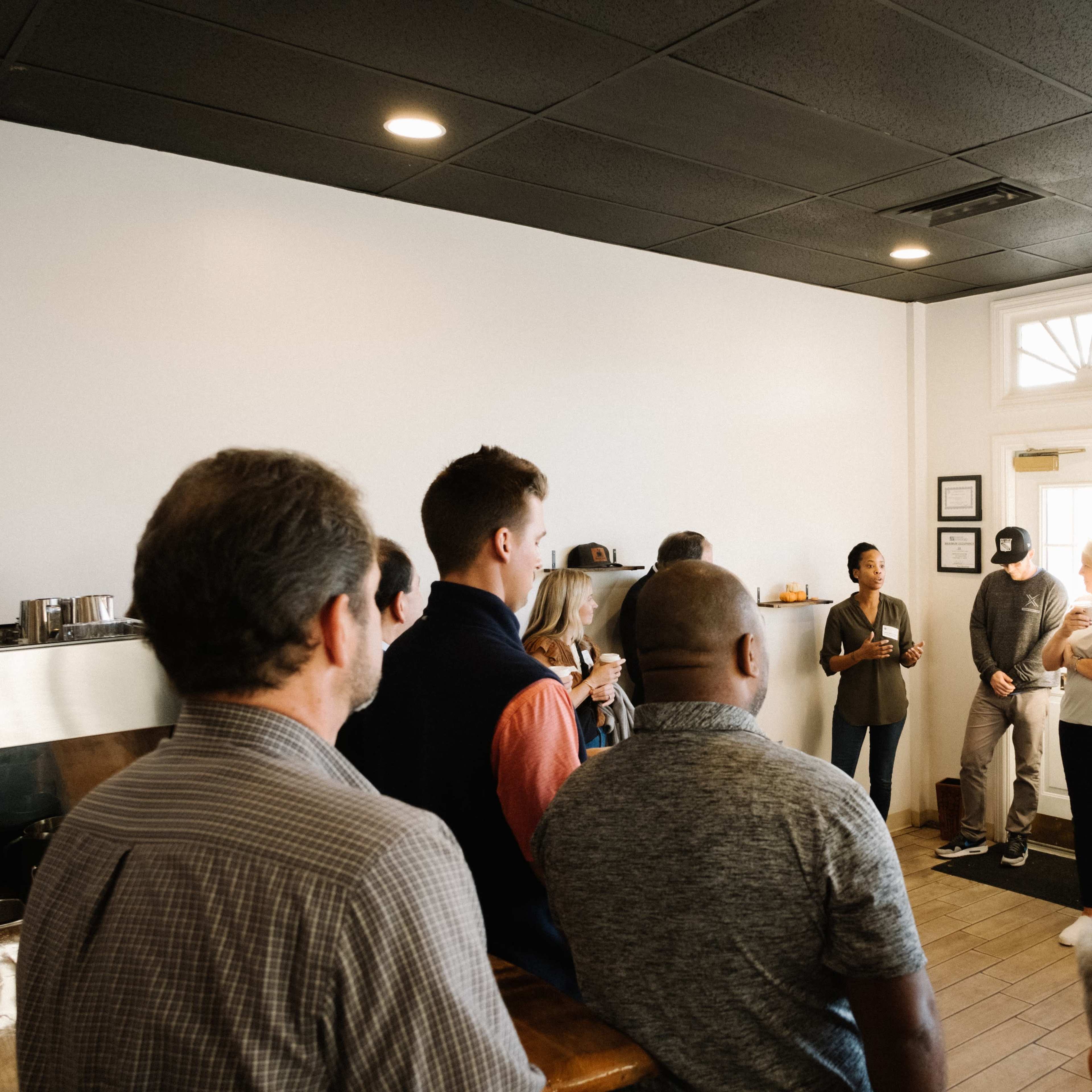 A group of people stands in a brightly lit room, engaging in conversation near a large window.