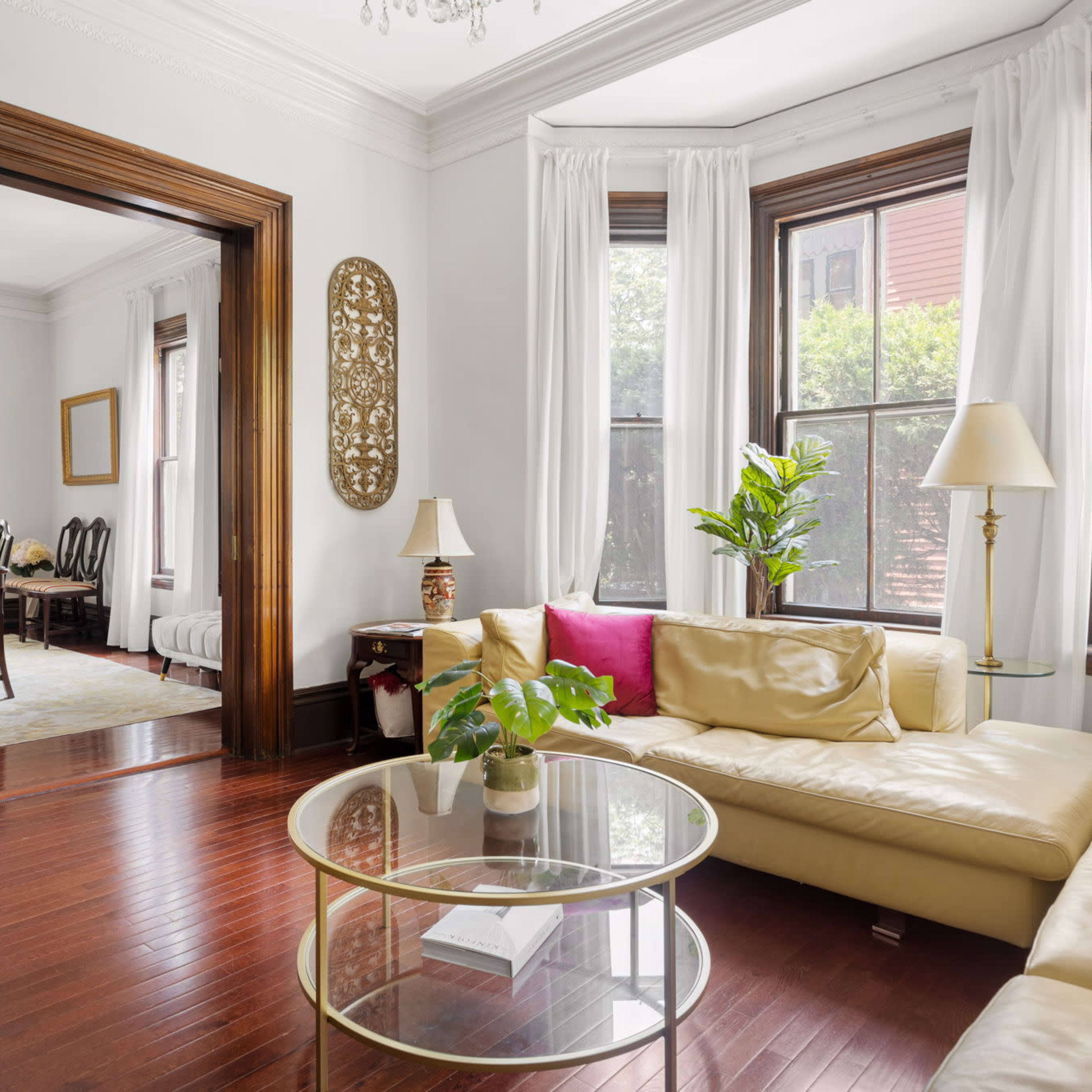 A living room features a beige sectional sofa, a round glass coffee table with a plant, and large windows allowing natural light, with a dining area visible in the background.