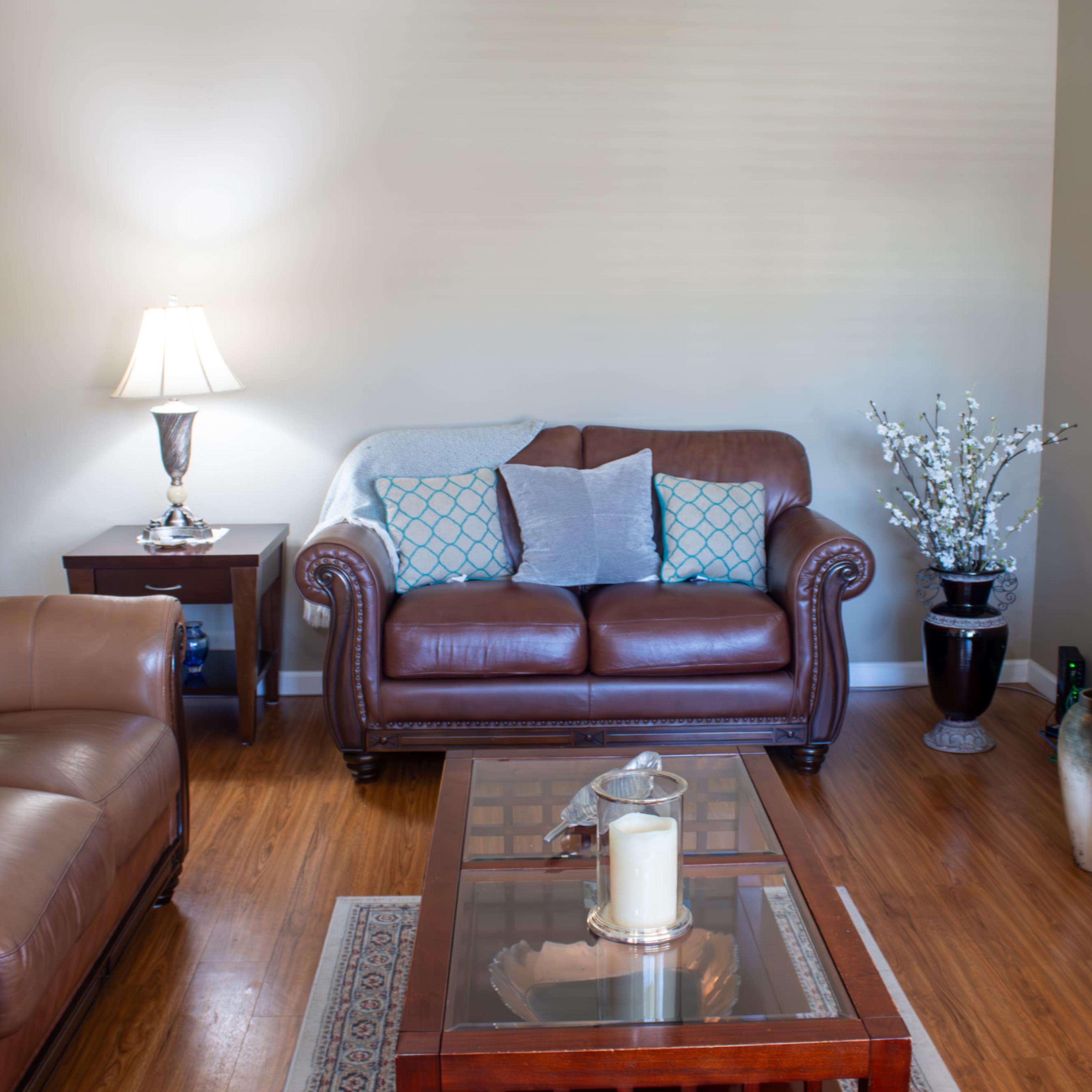 The living room features a brown leather sofa, a wooden coffee table with a candle, a side table with a lamp, and a television stand with a TV.