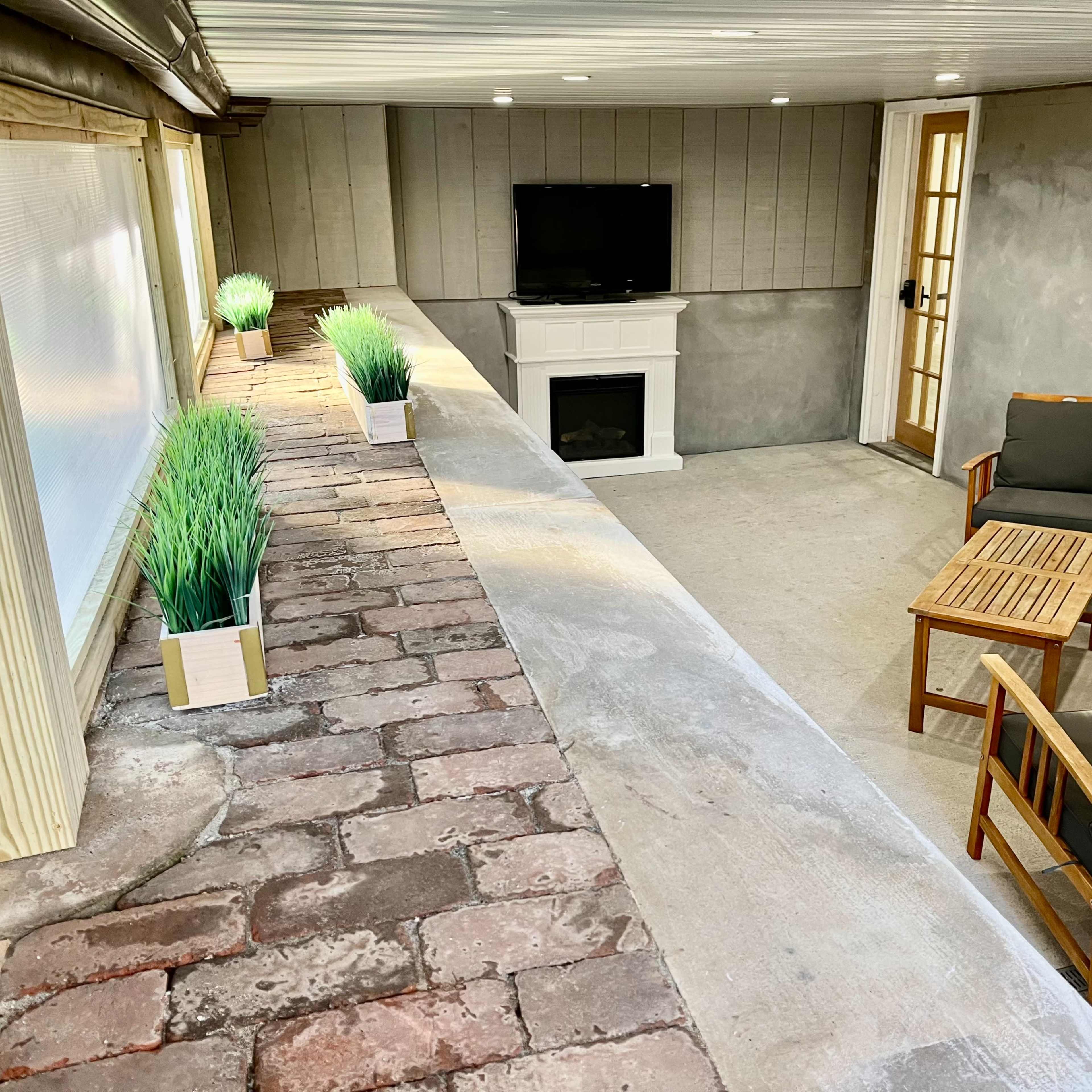 A cozy indoor space featuring a small seating area with two chairs, a wooden table, a fireplace, and a TV against a gray wall, enhanced by planters with greenery along the windowsill.