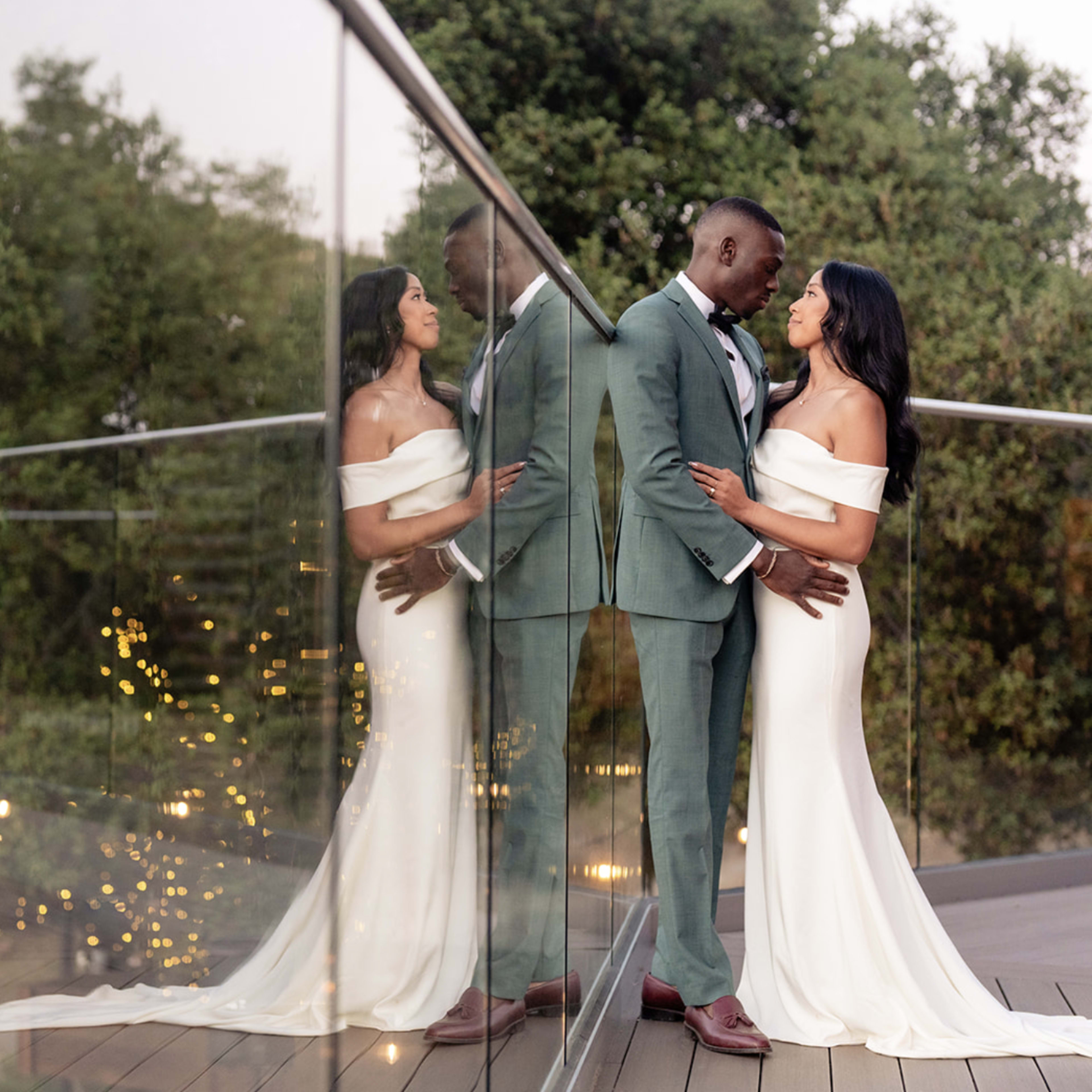 A couple stands close together on a balcony, with their reflections visible in the glass railing, surrounded by greenery and soft lights in the background.