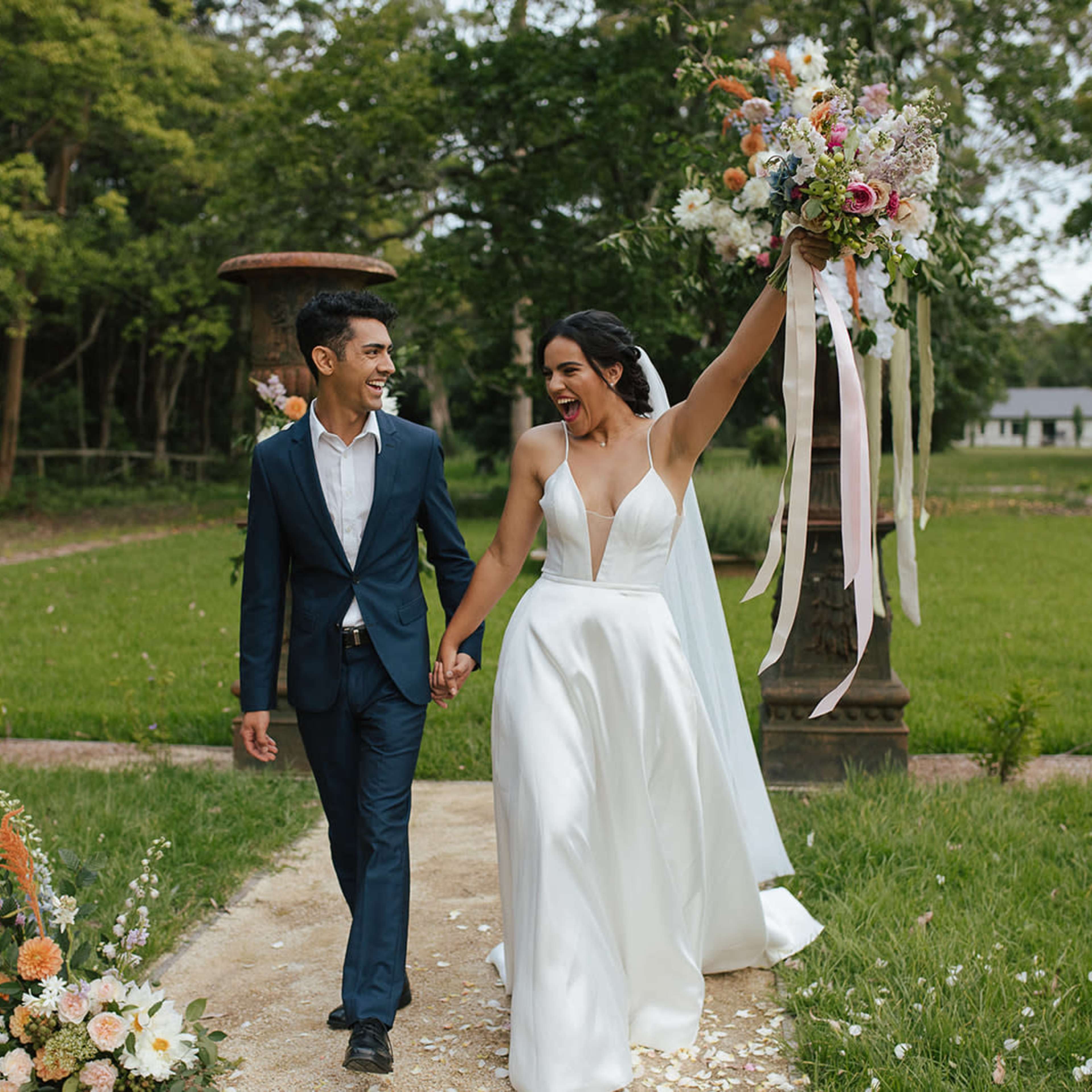 A newly married couple walks hand in hand down a garden pathway, with the bride joyfully raising her bouquet.