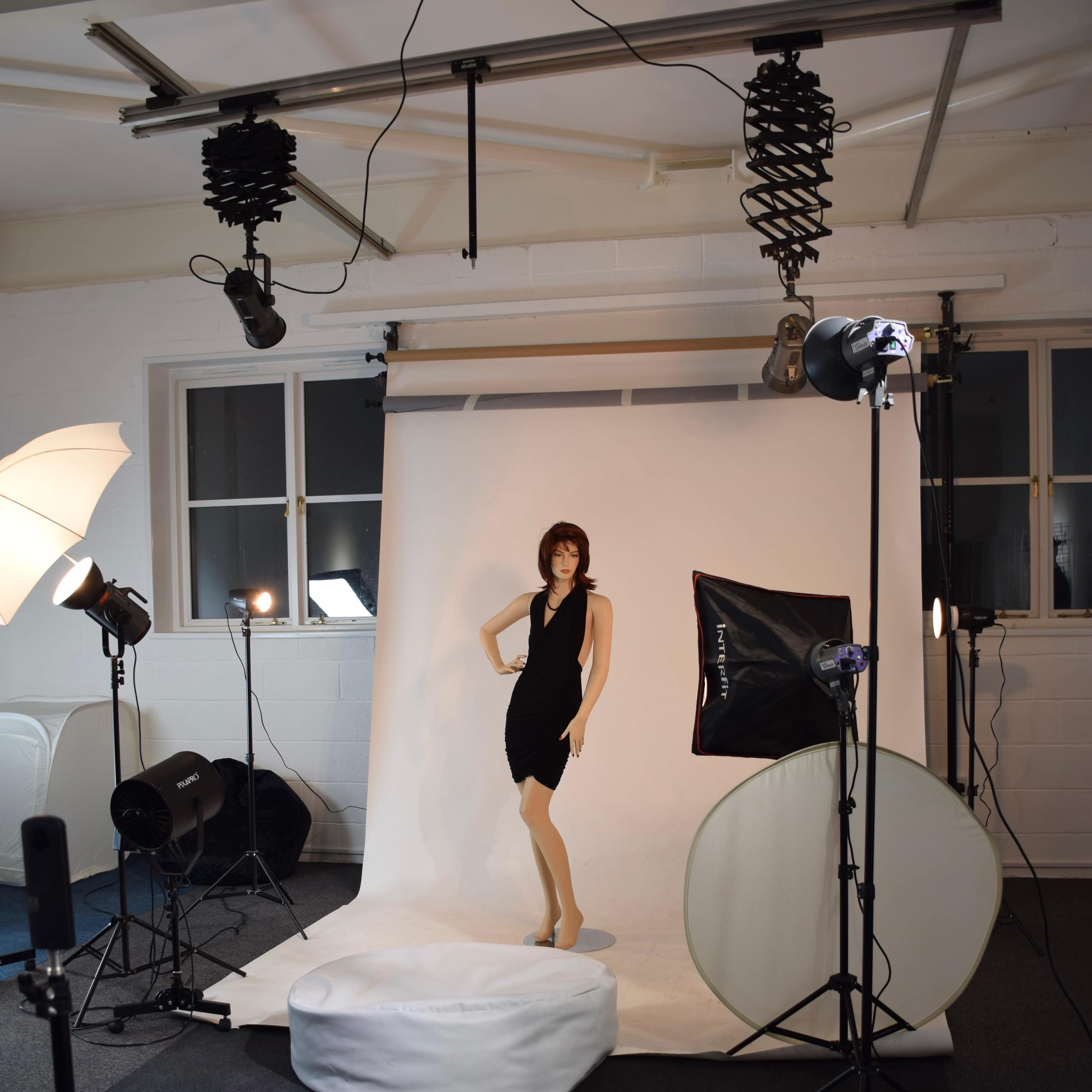 The scene features a mannequin dressed in a black dress, positioned on a circular platform in a photography studio surrounded by various lighting equipment.