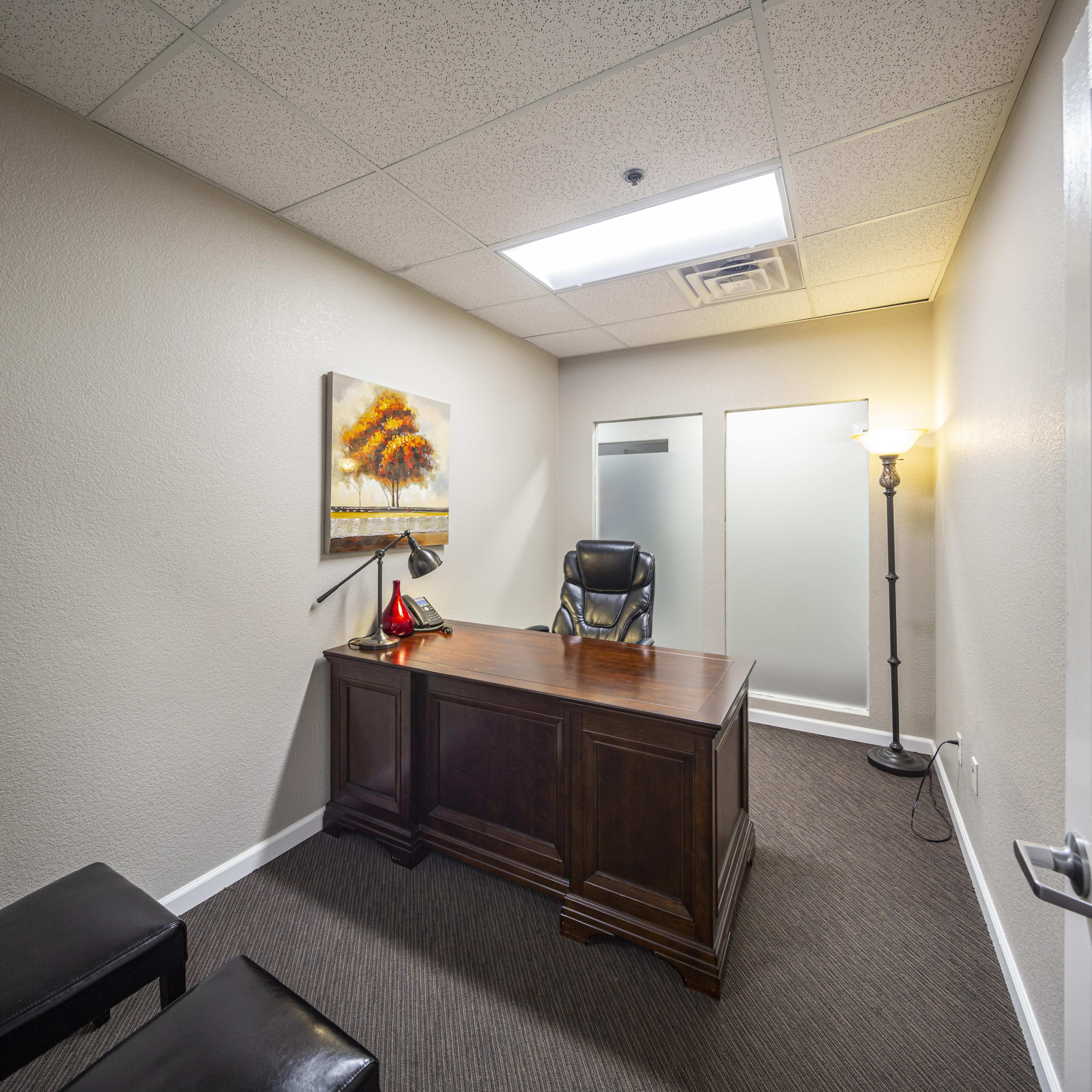 A small office features a dark wooden desk with a chair, a painting on the wall, and a floor lamp in the corner.