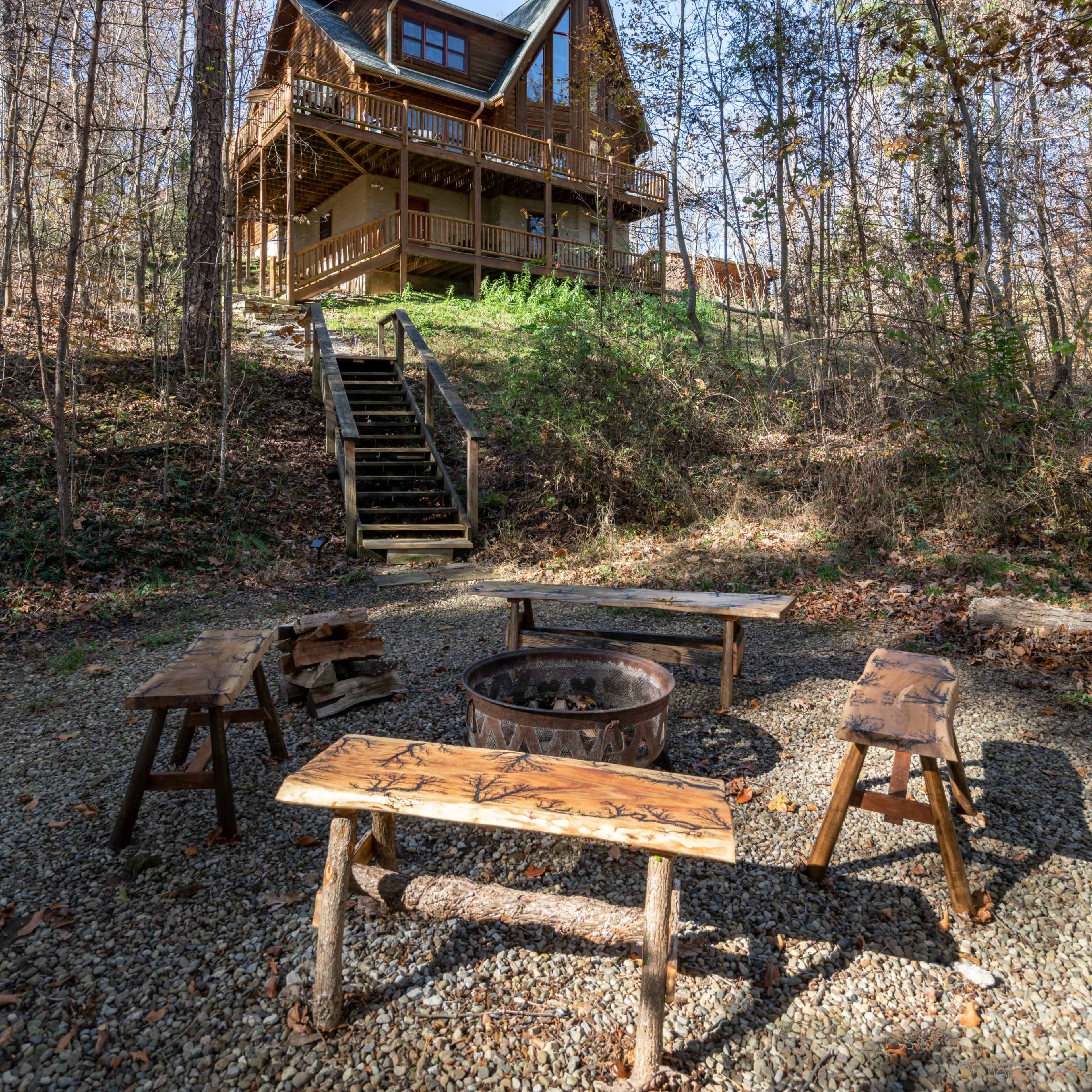 A wooden cabin with a steep roof stands on a hillside, surrounded by trees, with a fire pit and benches arranged in front of it.