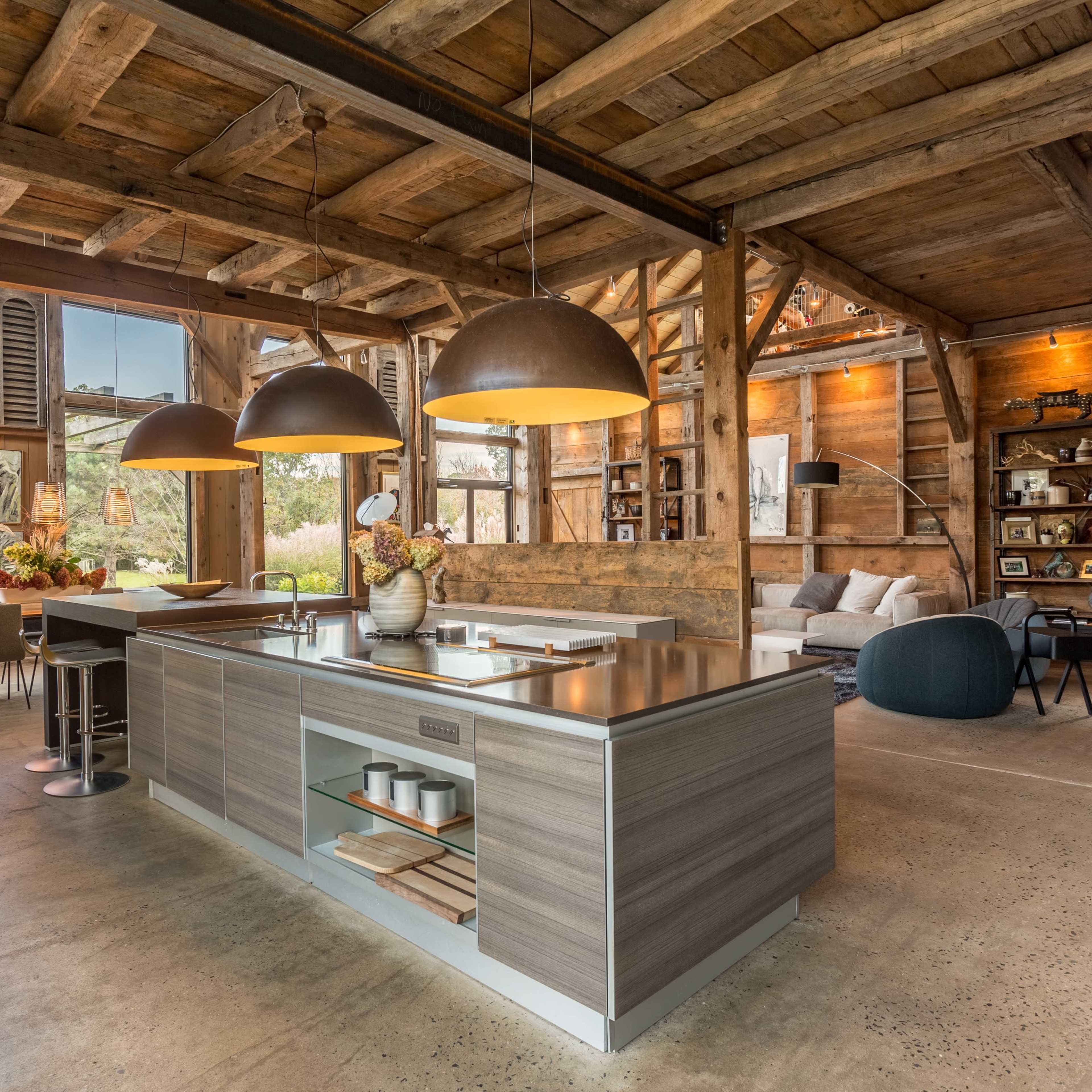 A modern kitchen with a central island, wooden beams, and large pendant lights, set in a rustic interior with large windows.