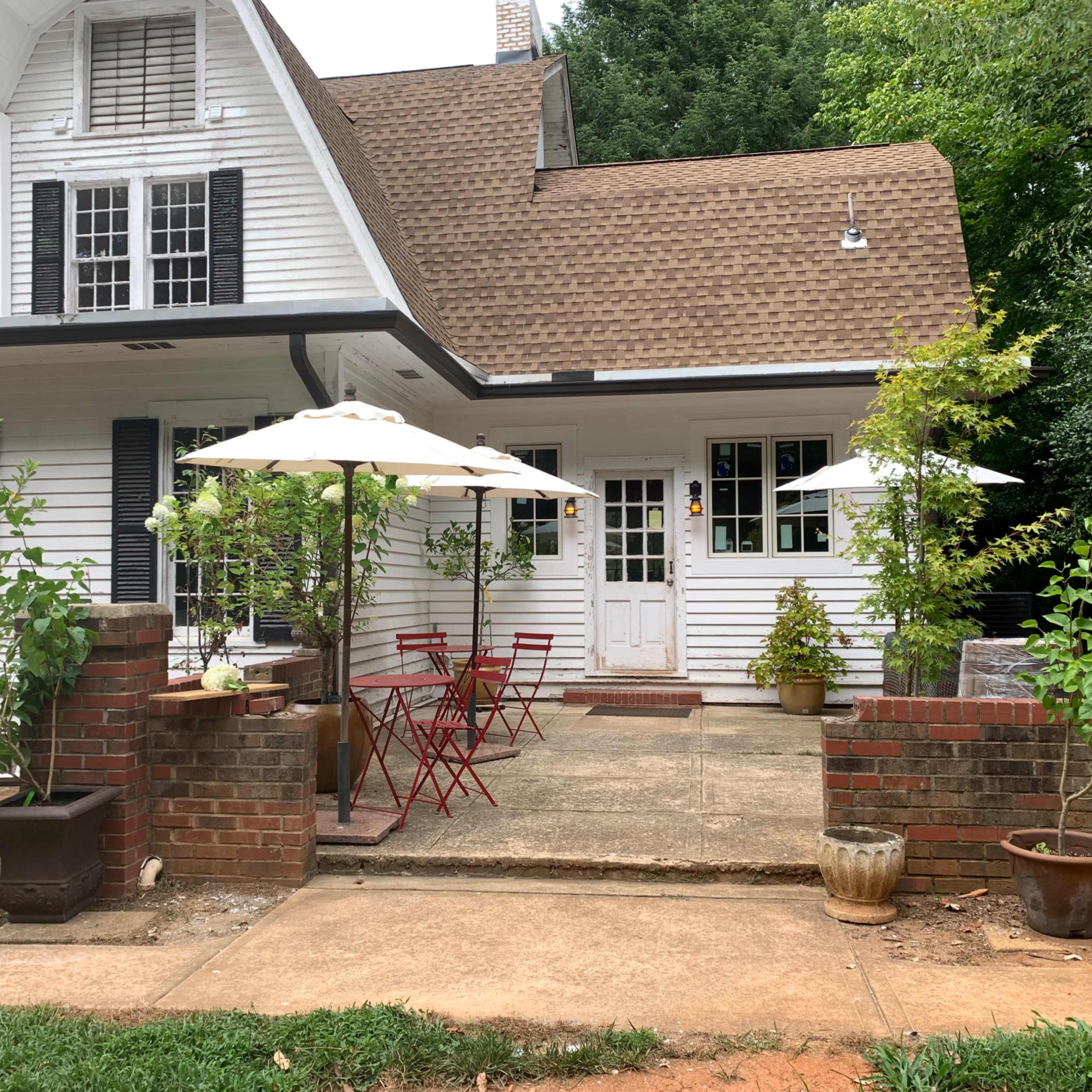 A charming house with a white exterior features a small patio area with red metal chairs, a table, and large umbrellas, surrounded by potted plants.