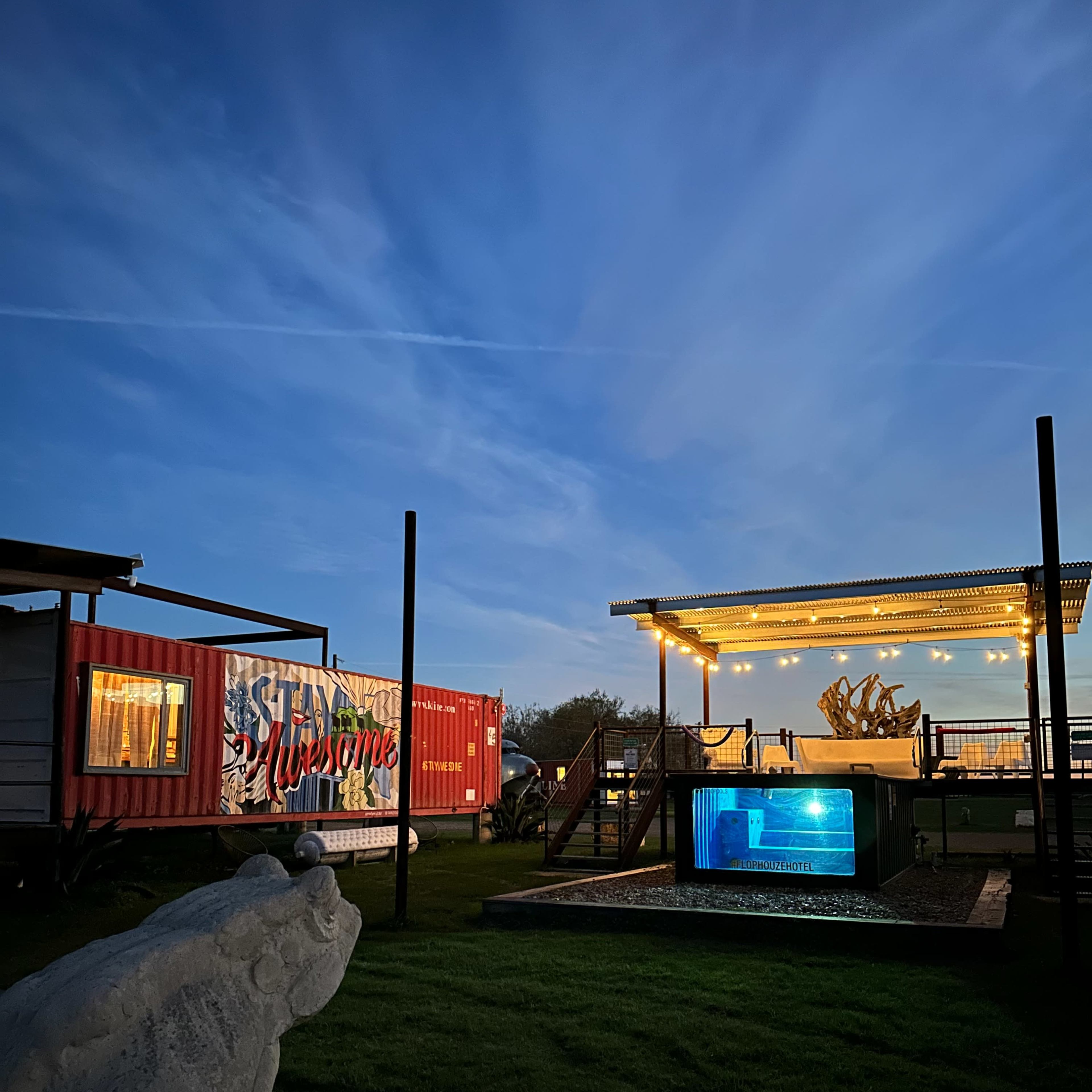 A colorful shipping container marked "Welcome," a well-lit patio with decorative lights, and a stone frog sculpture in a grassy area during twilight.