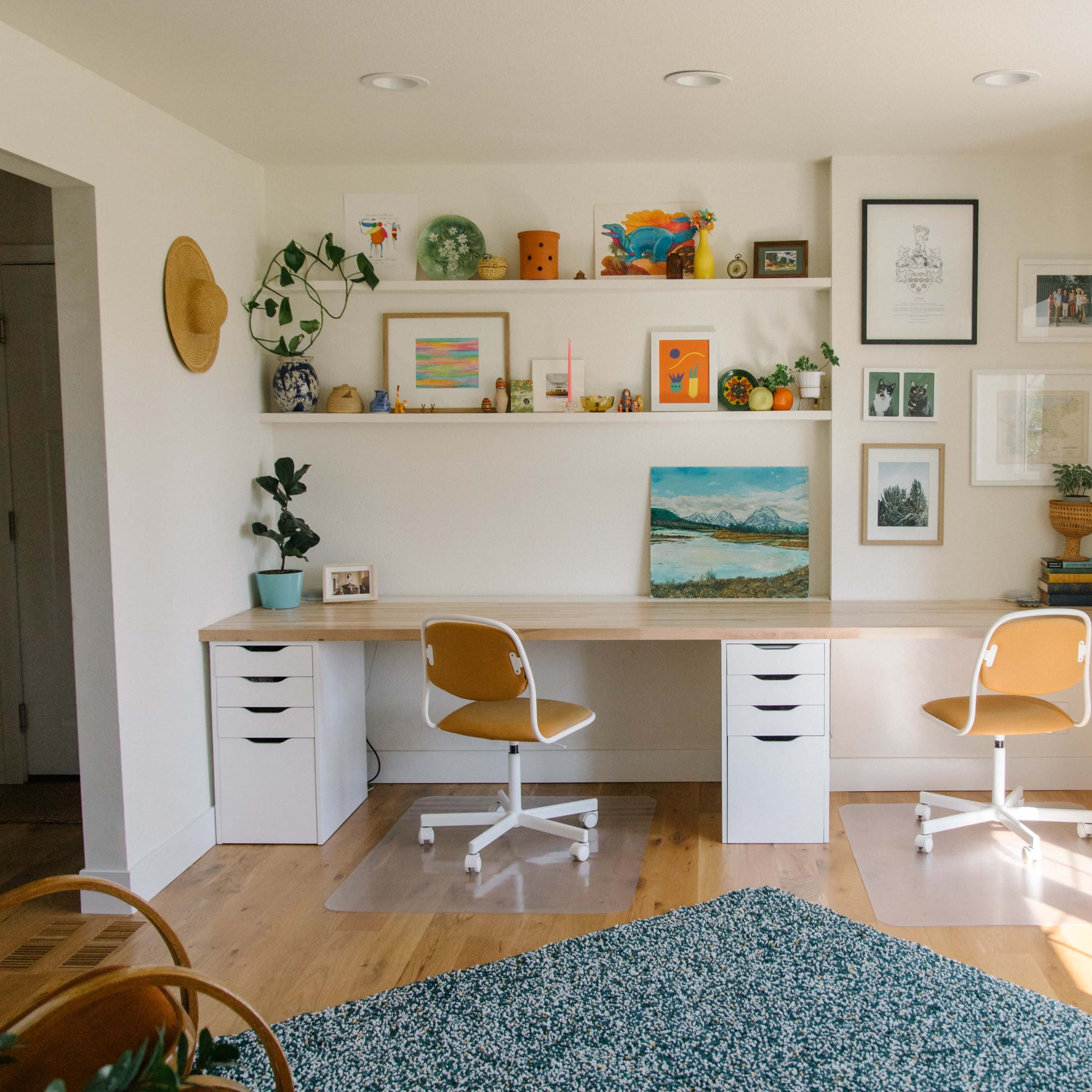 A bright home office with two chairs at a long desk, surrounded by plants and wall art on shelves.