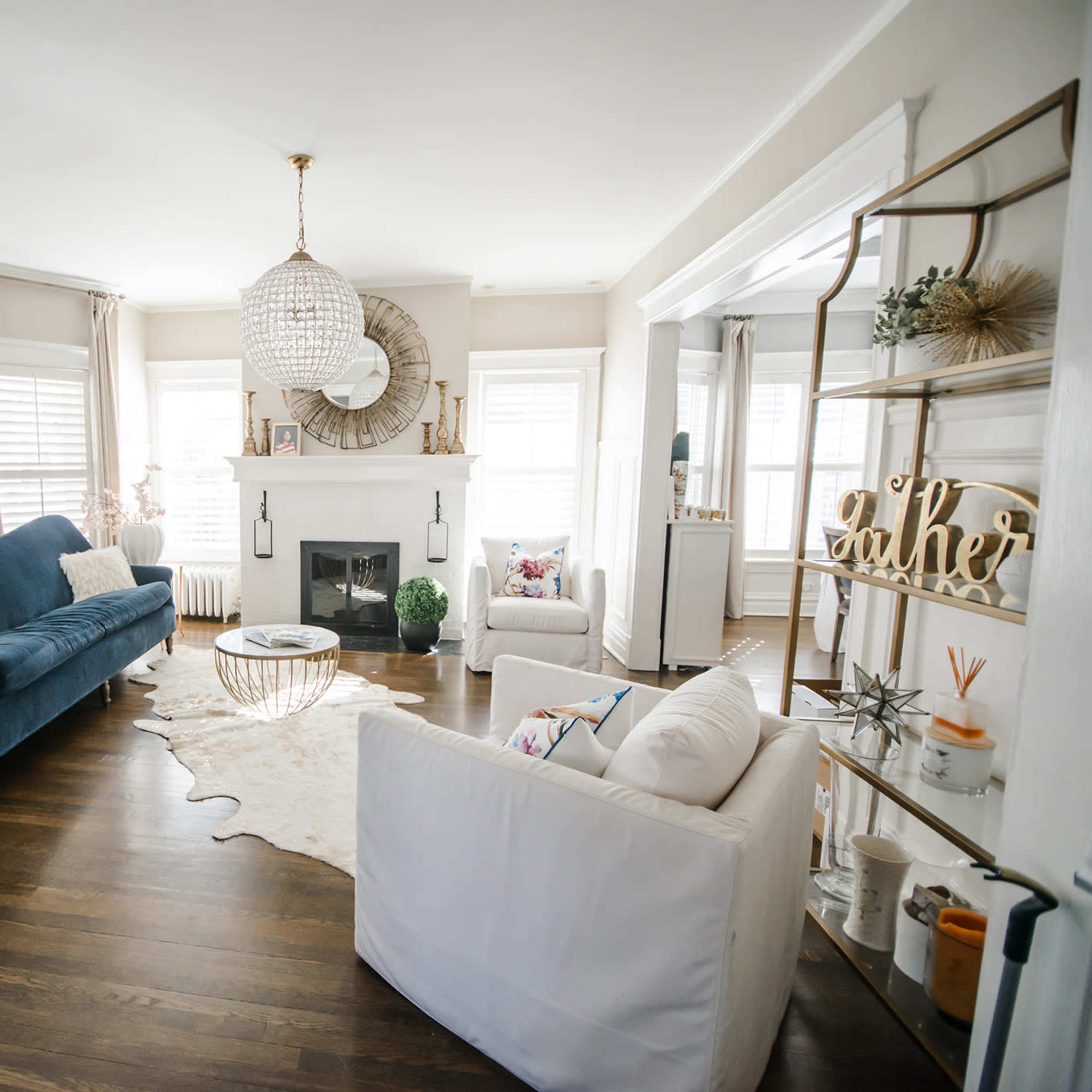 A modern living room features a blue sofa, white chairs, a round coffee table, a decorative rug, and a chandelier.