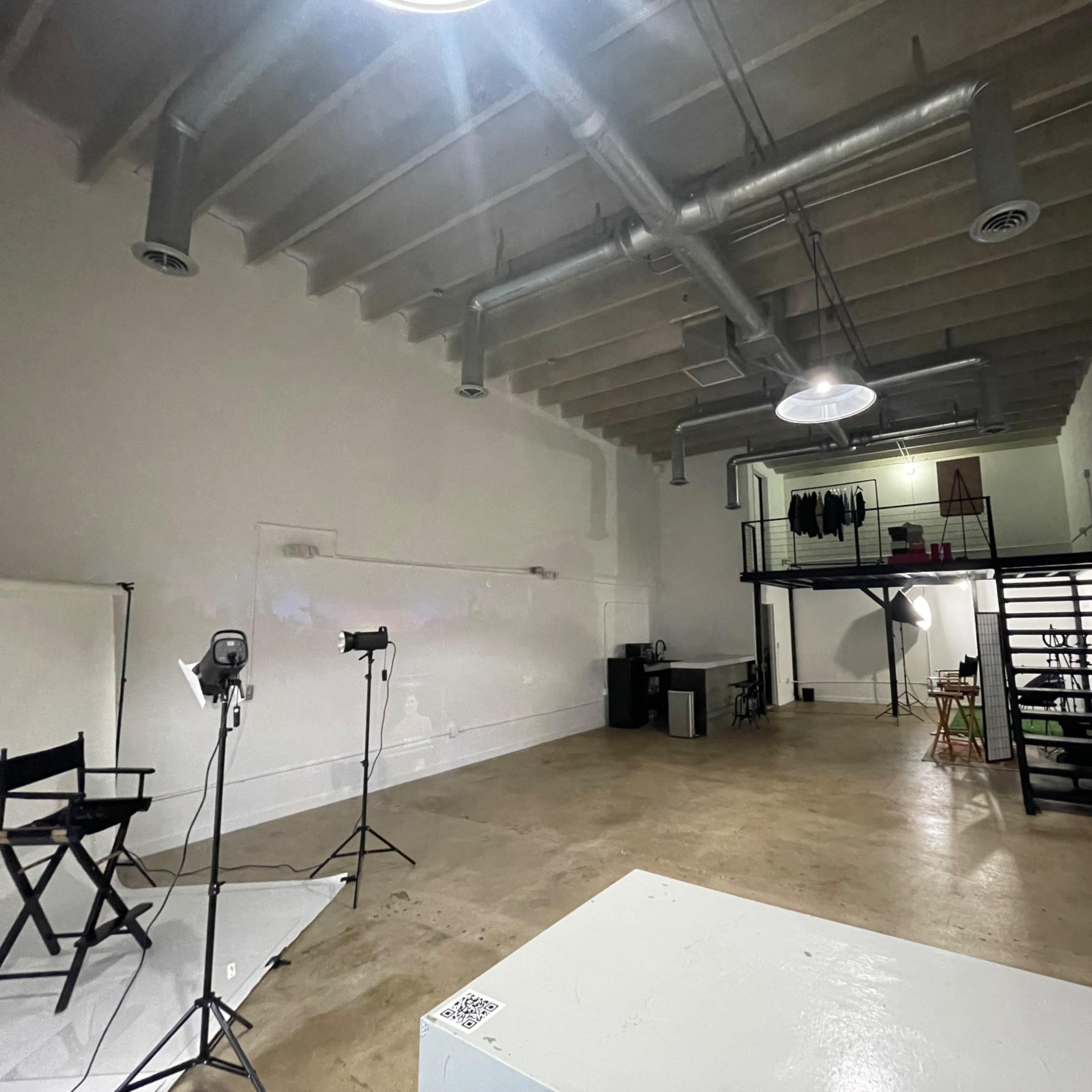 A spacious, empty studio with a wooden floor, high ceilings, and a staircase in the back.