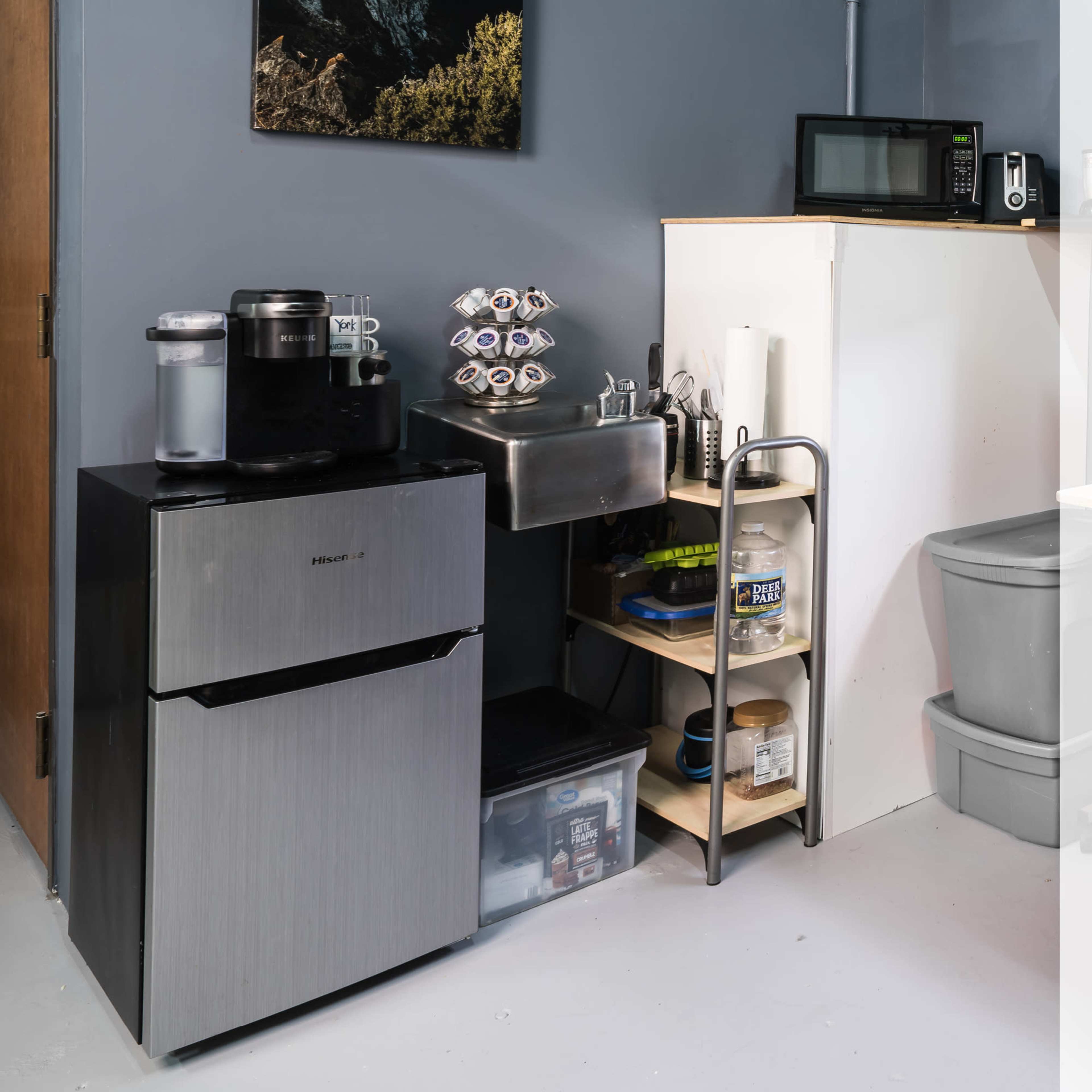A kitchenette area with a refrigerator, coffee machine, sink, and shelves containing various items in a small space.
