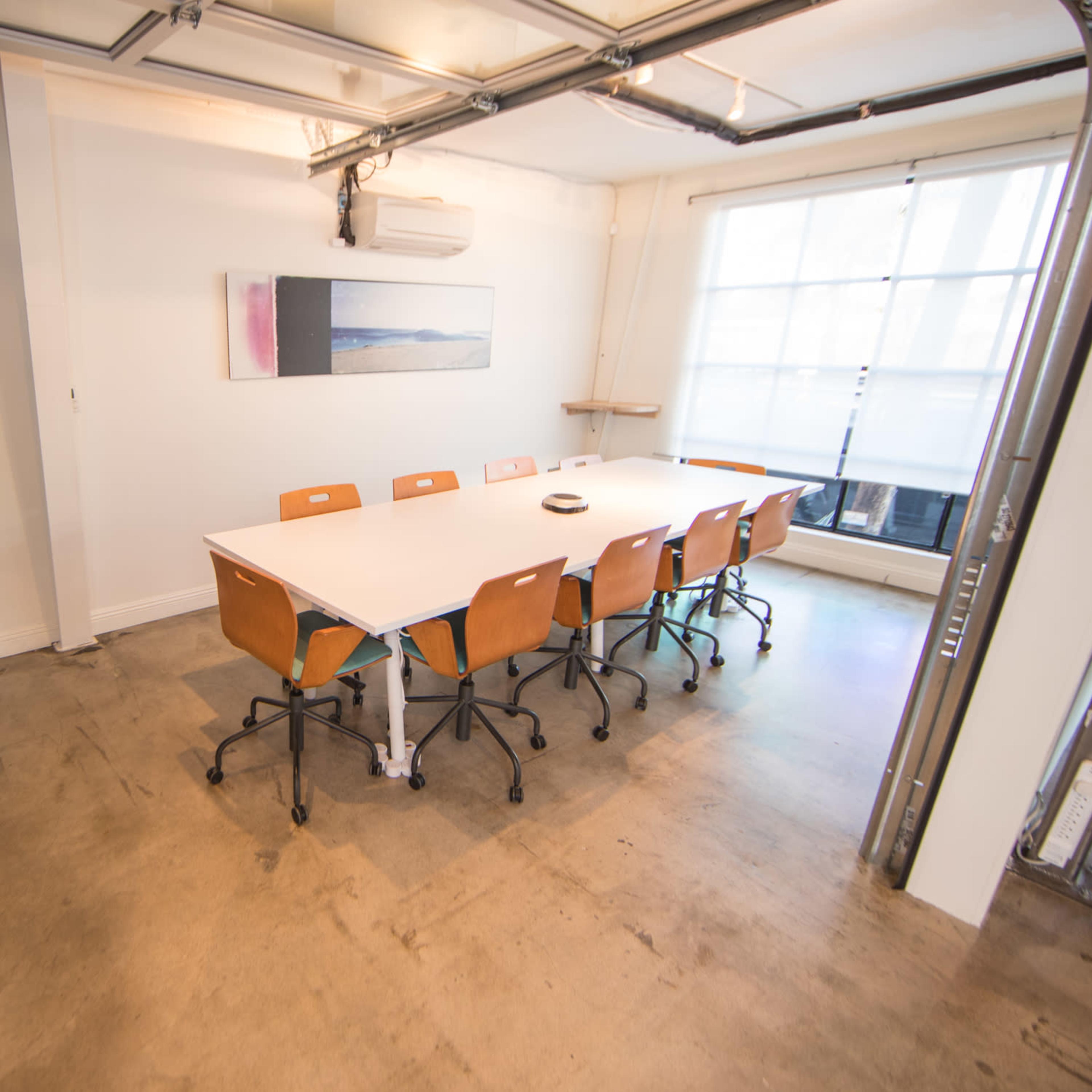 A minimalistic meeting room features a large white table surrounded by orange chairs, with a television mounted on the wall and a window letting in natural light.
