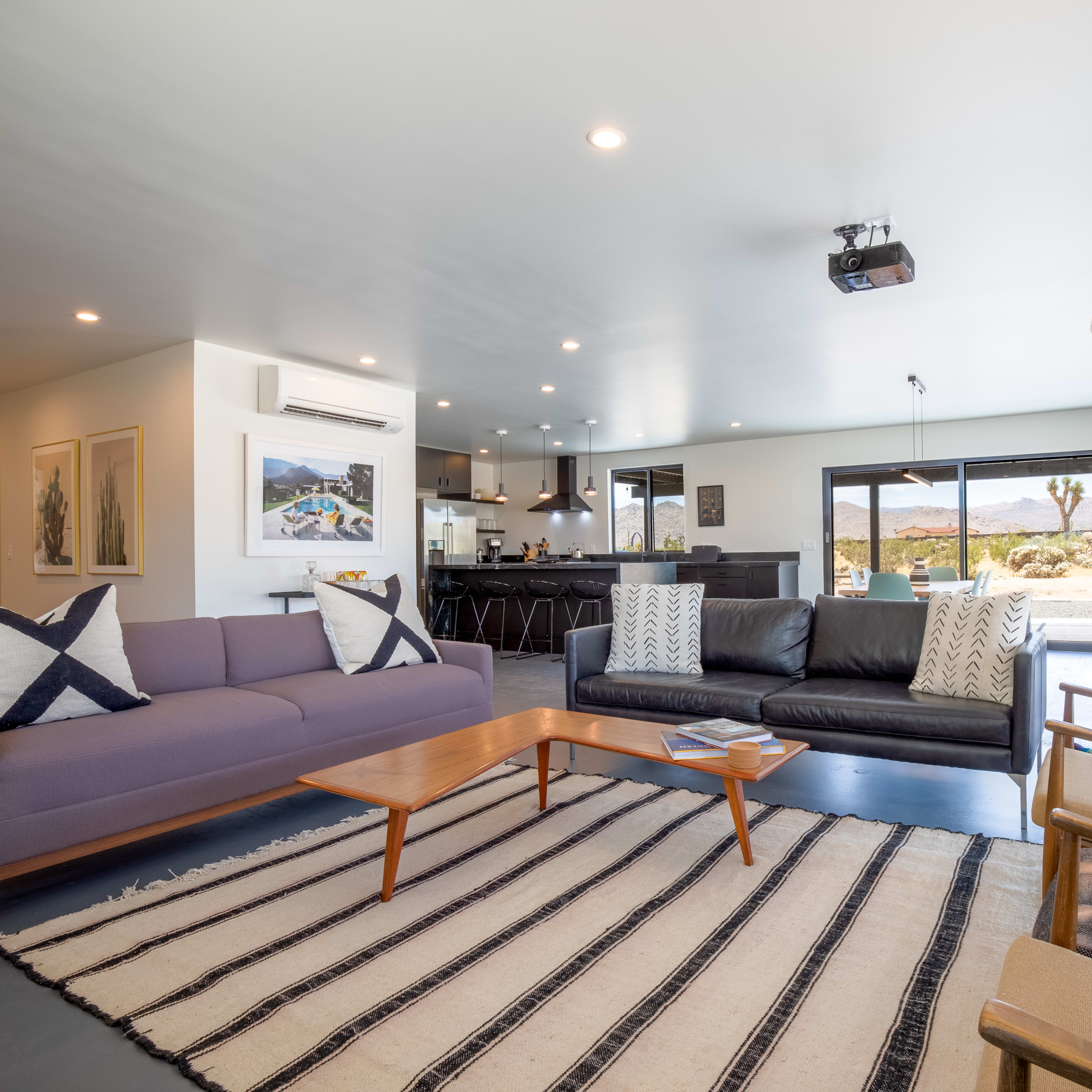 The living room features a mix of modern furniture, including a purple sofa and a black leather couch, arranged around a wooden coffee table on a striped area rug, with large windows offering a view of the desert landscape.