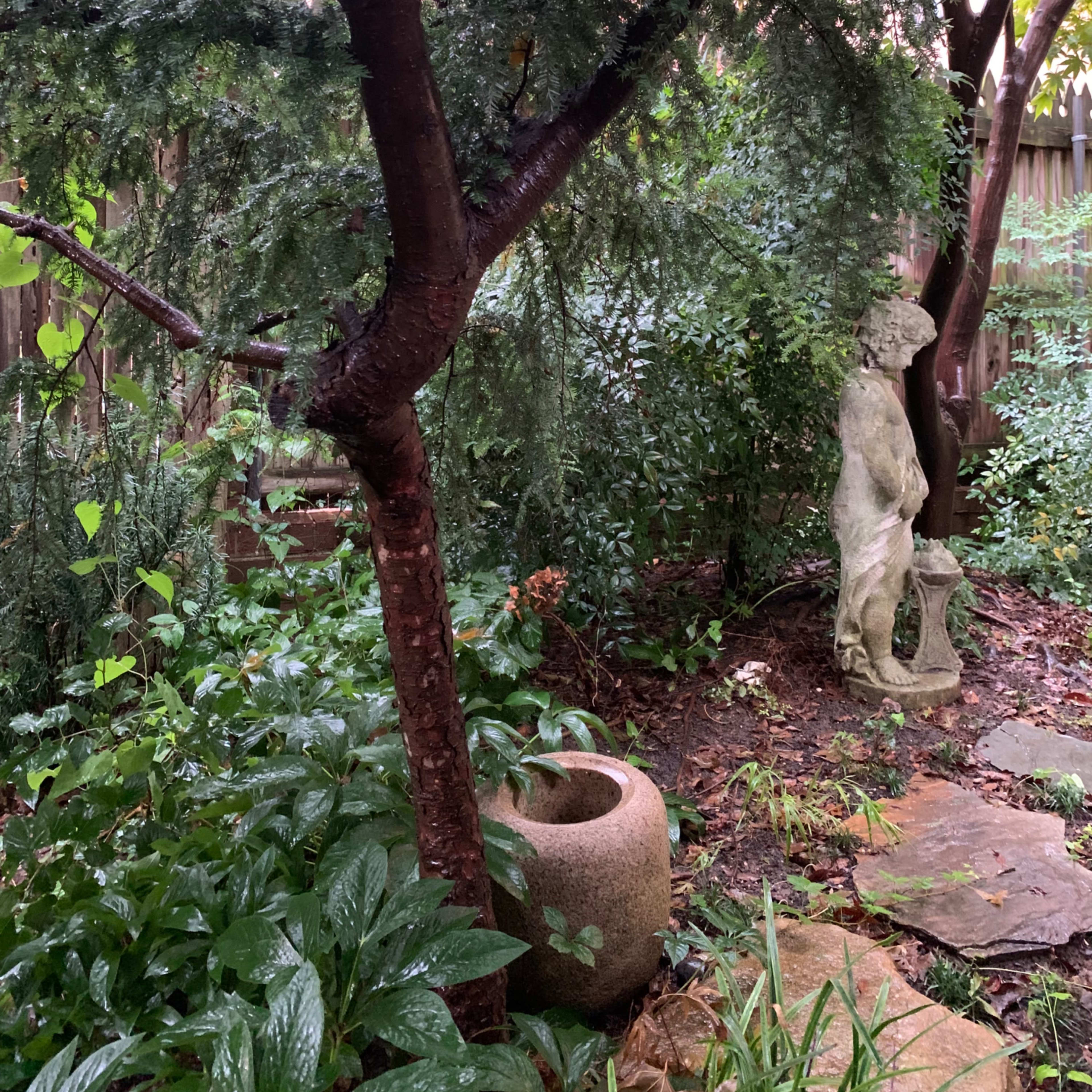 The scene features a lush garden with a stone statue partially obscured by greenery, a textured tree trunk, and a round stone planter.