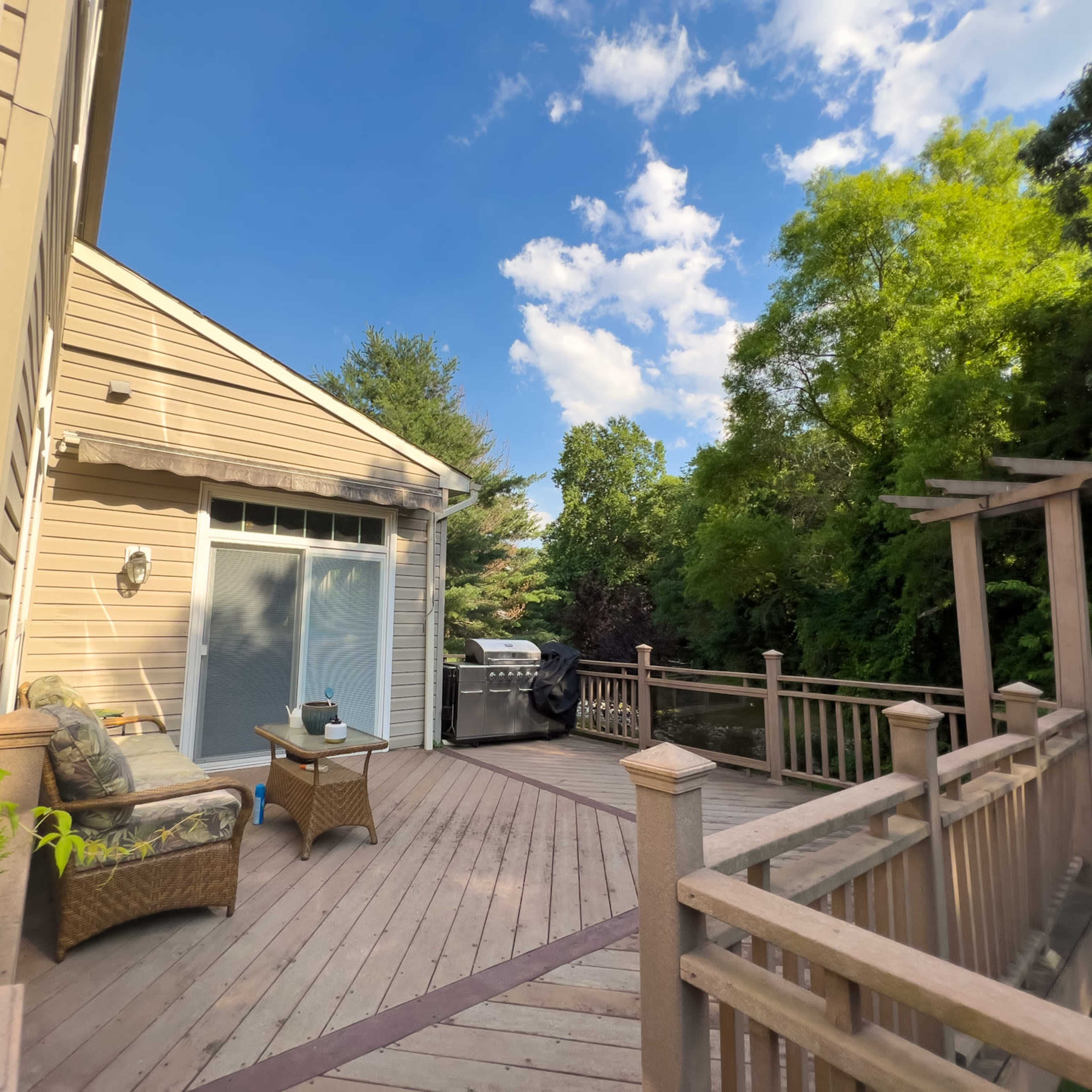 A wooden deck with outdoor furniture and a grill, surrounded by greenery and under a partly cloudy sky.