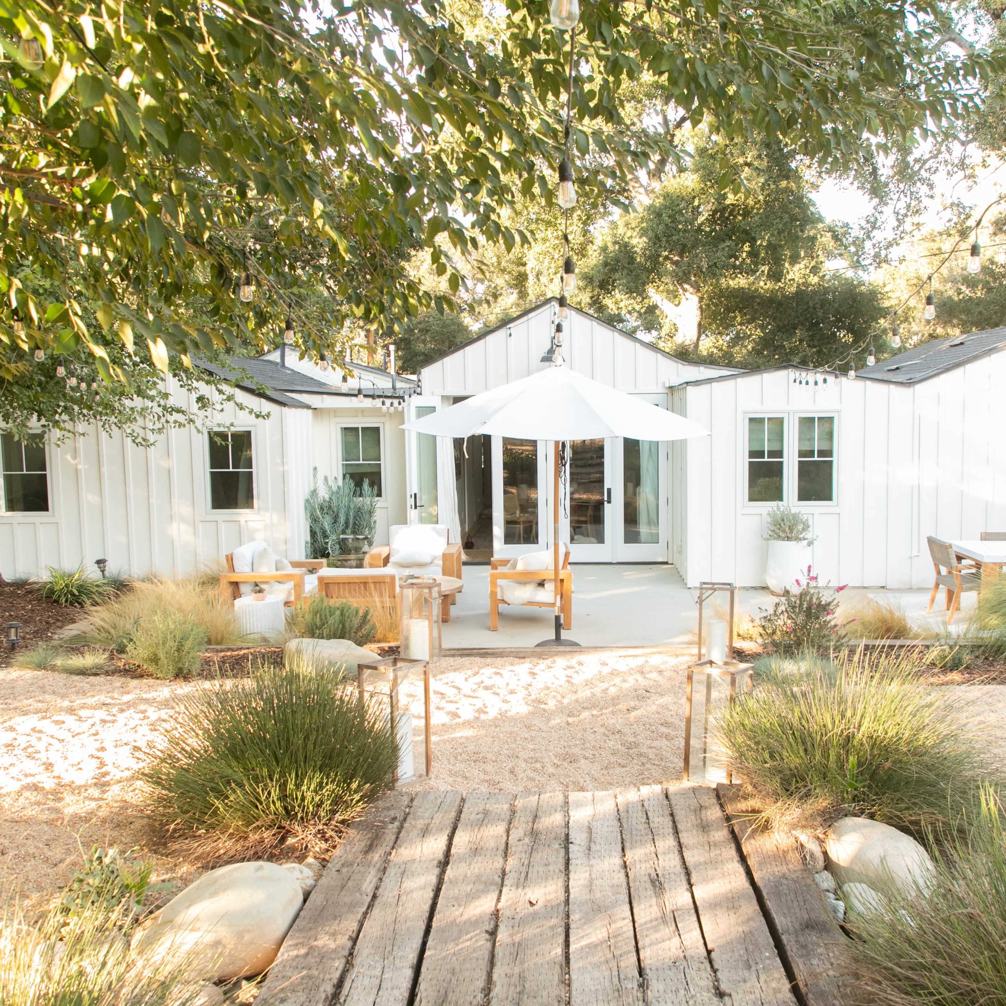 A rustic white house with a shaded patio and outdoor seating is surrounded by landscaped gardens and a pathway made of wooden planks.