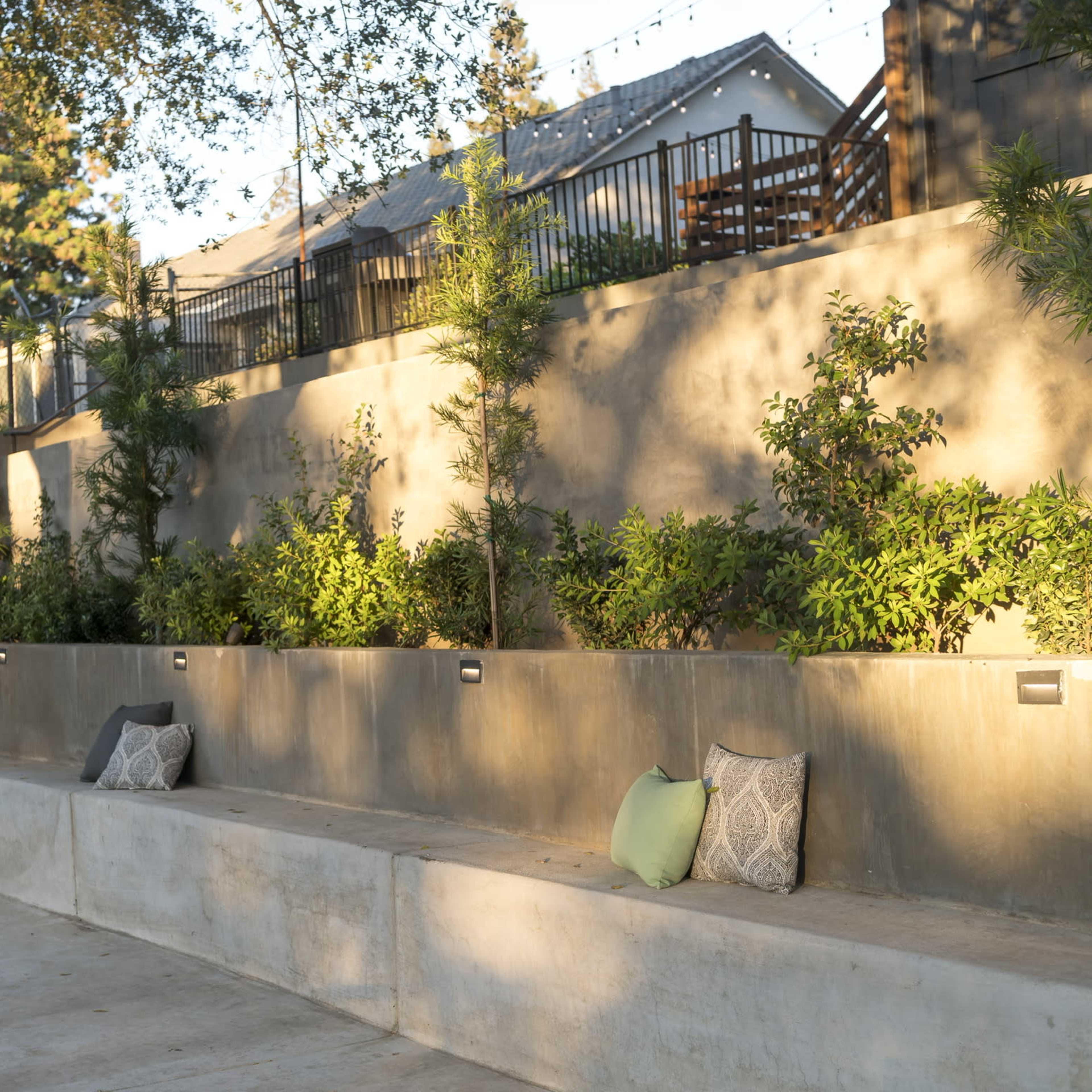 A concrete seating area with decorative pillows, bordered by greenery and under trees, with residential structures visible in the background.