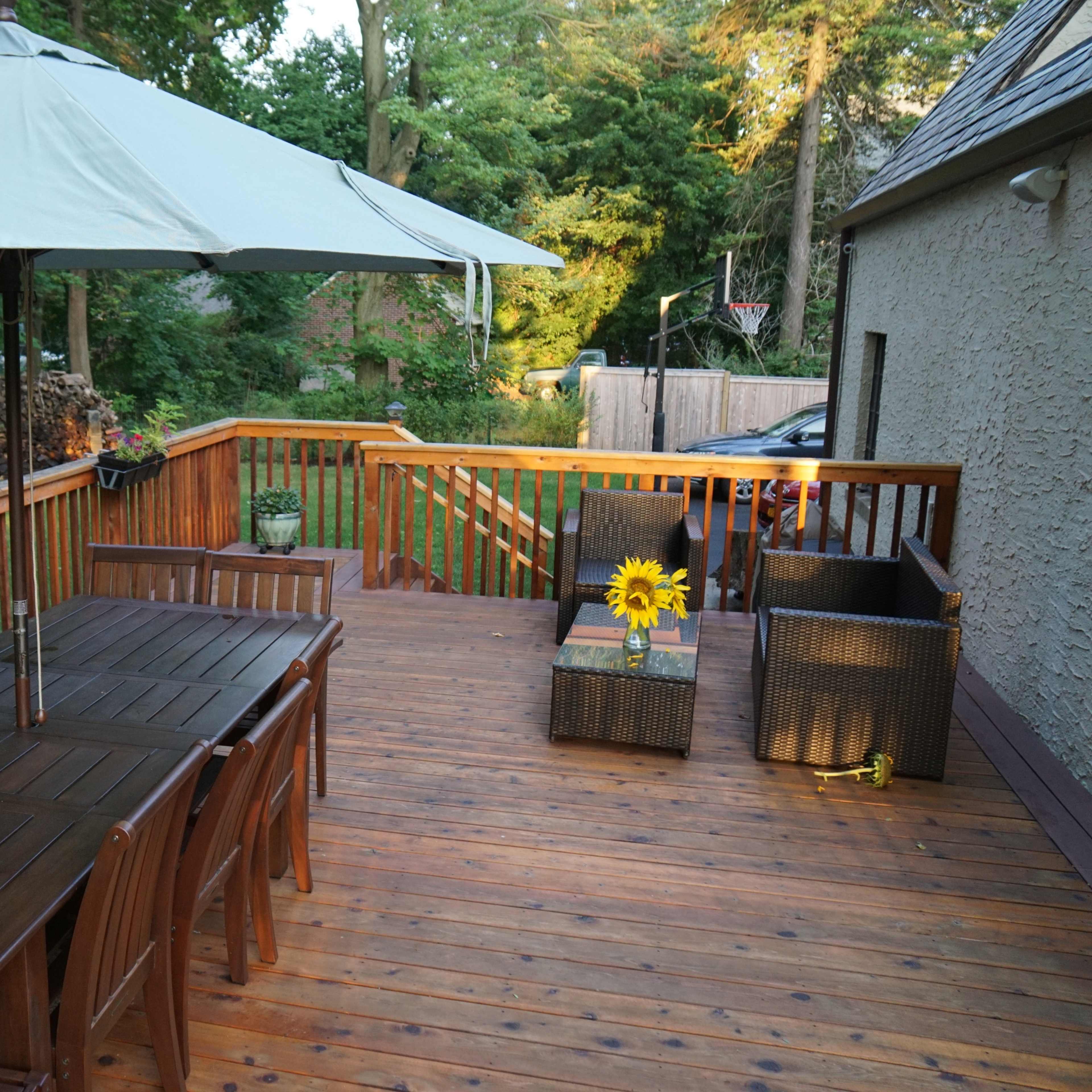 A wooden deck features a large dining table with an umbrella, surrounded by potted plants and wicker furniture.