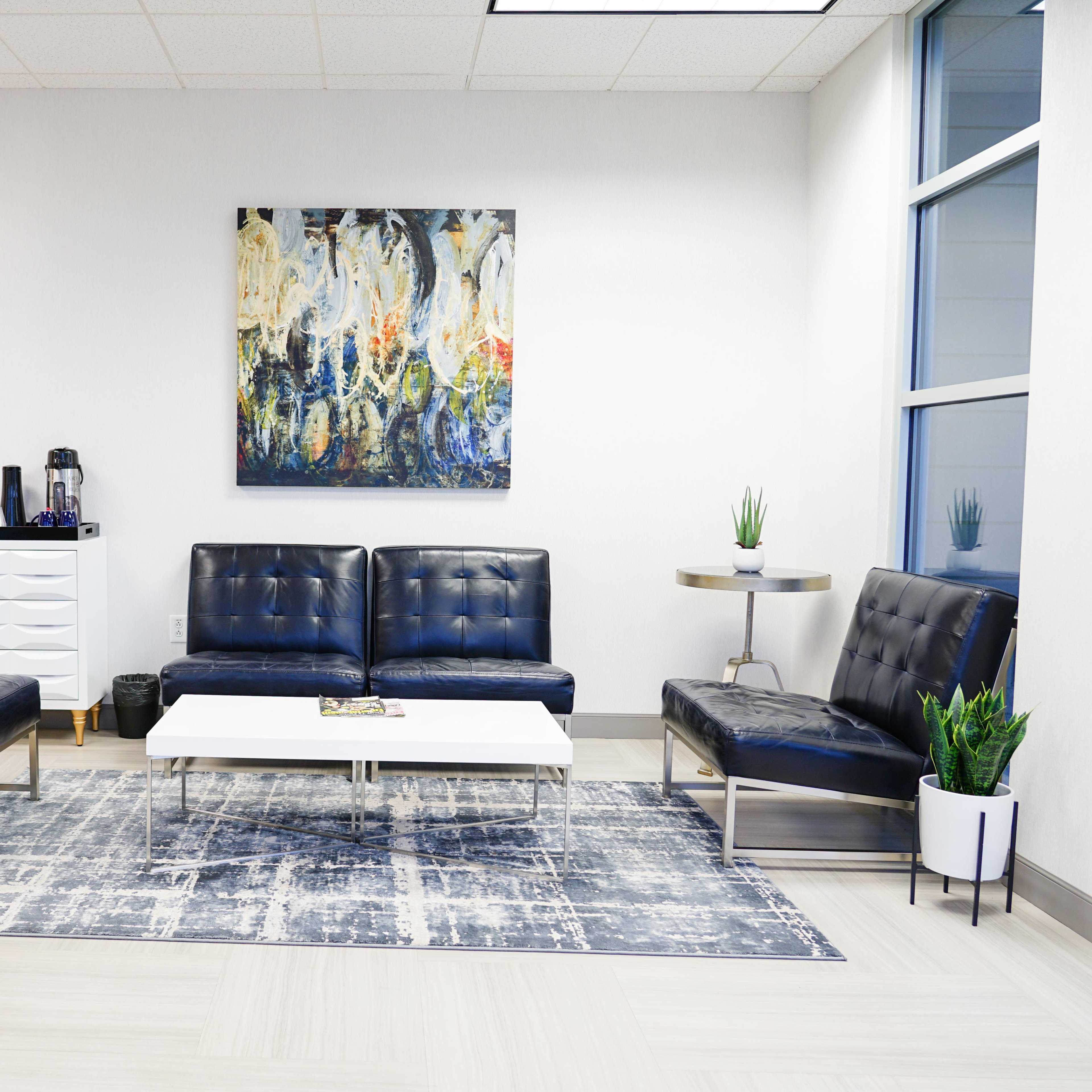 A modern waiting area featuring two black leather couches, two matching chairs, a coffee table, and a decorative wall art piece, all set on a patterned area rug.