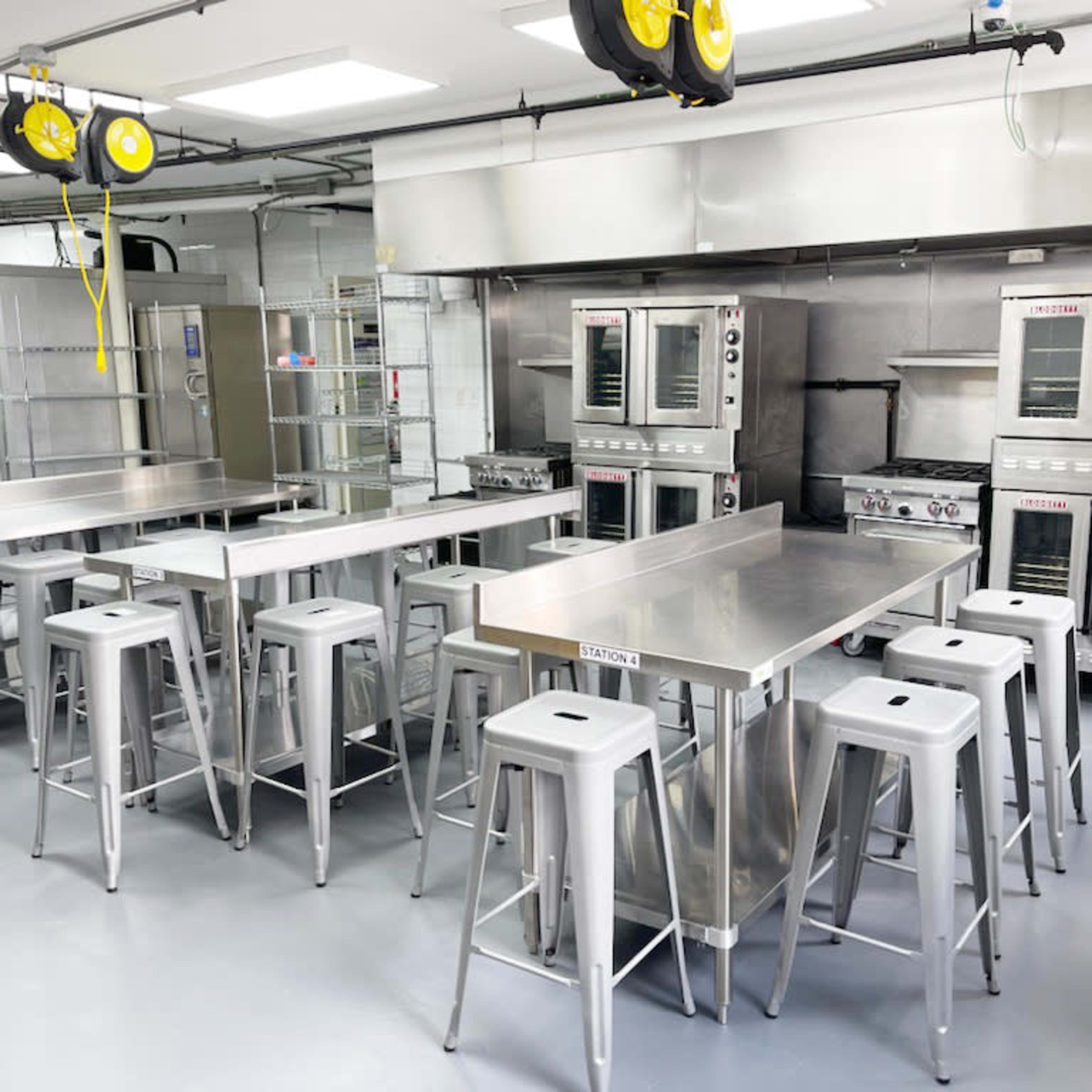 A commercial kitchen is equipped with stainless steel tables, high stools, and multiple ovens and cooking appliances.