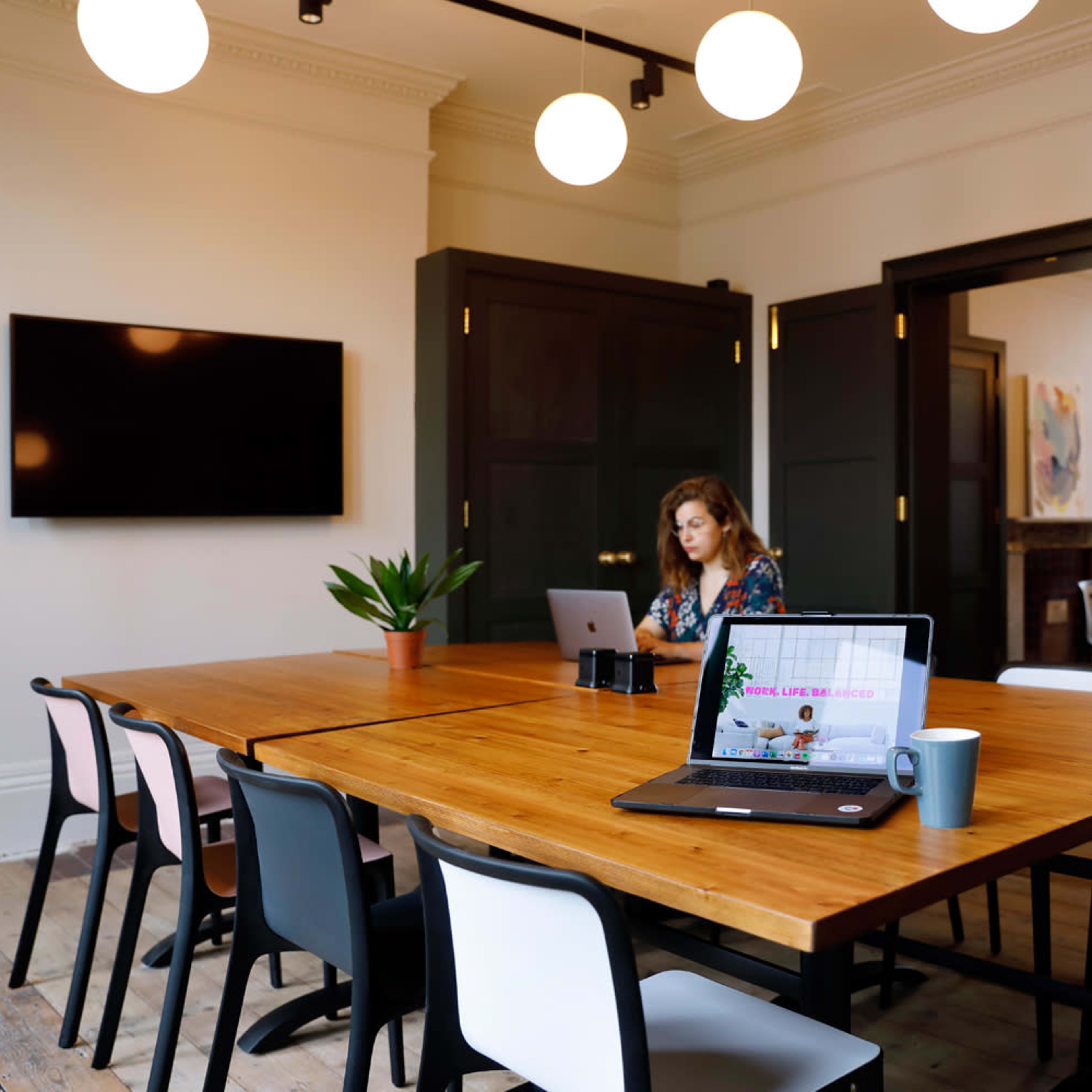 A woman works on a laptop at a wooden table in a conference room with round lights and green plants.