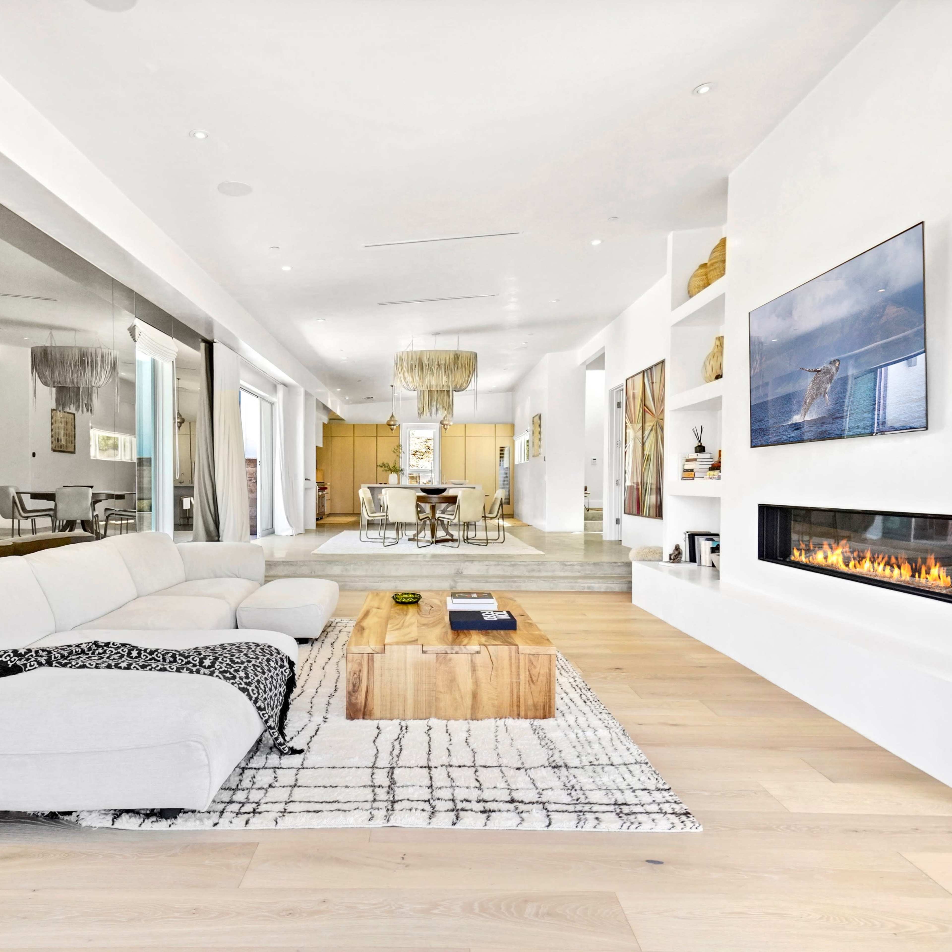 A modern living room featuring a light-colored sectional sofa, a wooden coffee table, a wall-mounted television, and a linear fireplace, all set against a backdrop of white walls and large windows.