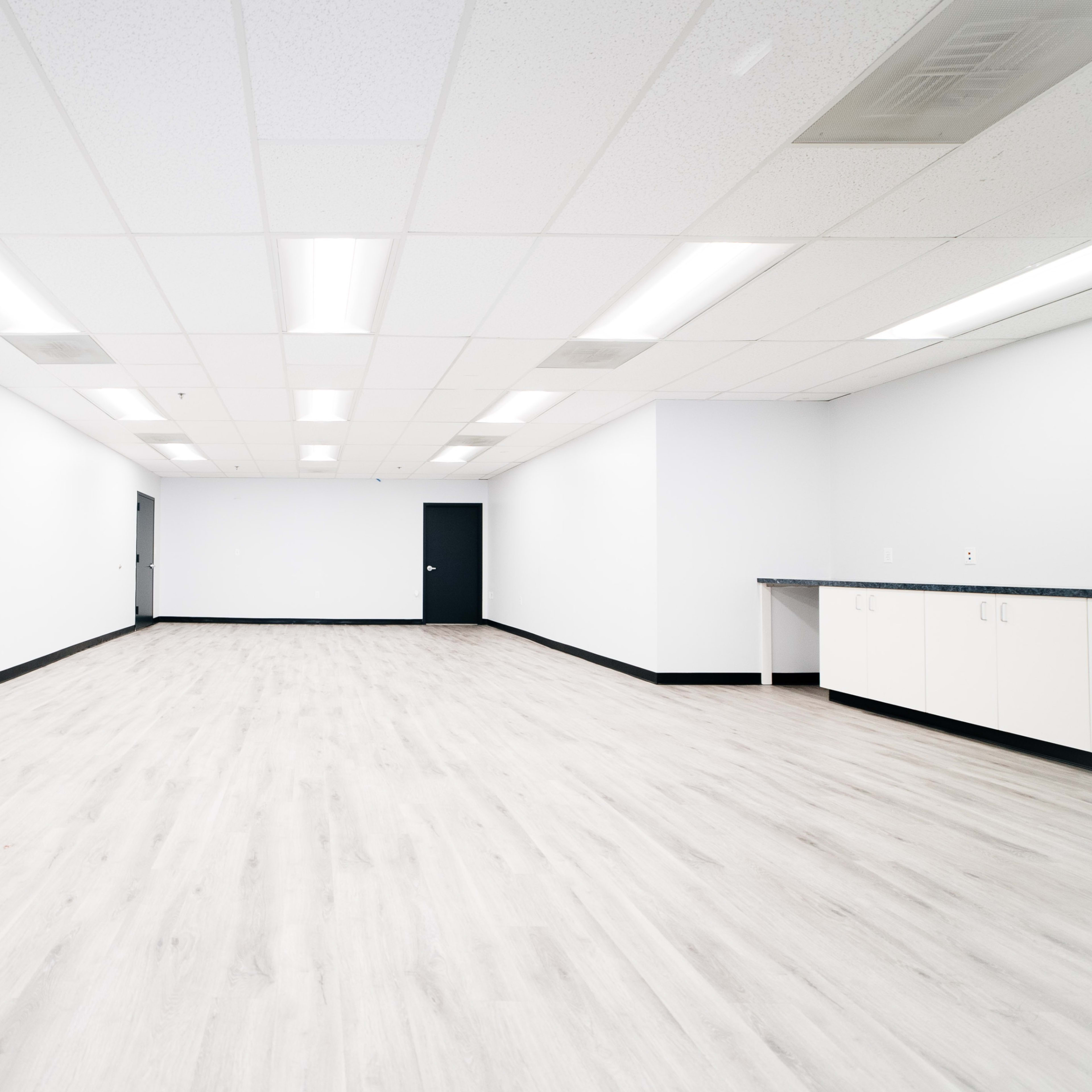 A brightly lit, empty room with light-colored walls, a wooden floor, and a dark door and counter along one side.
