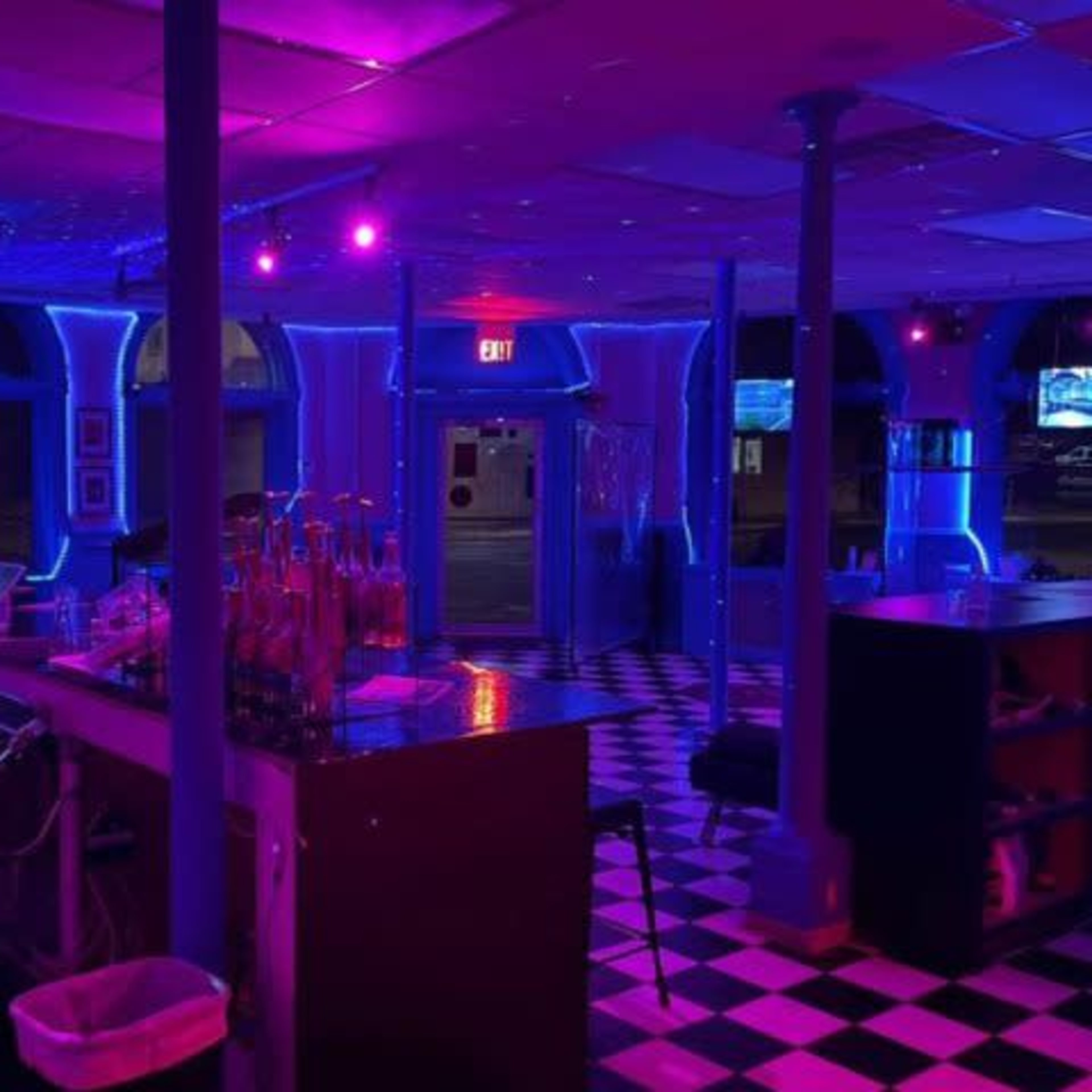 A brightly lit bar features purple and blue neon lights, with a checkered floor and rows of bottles behind the counter.