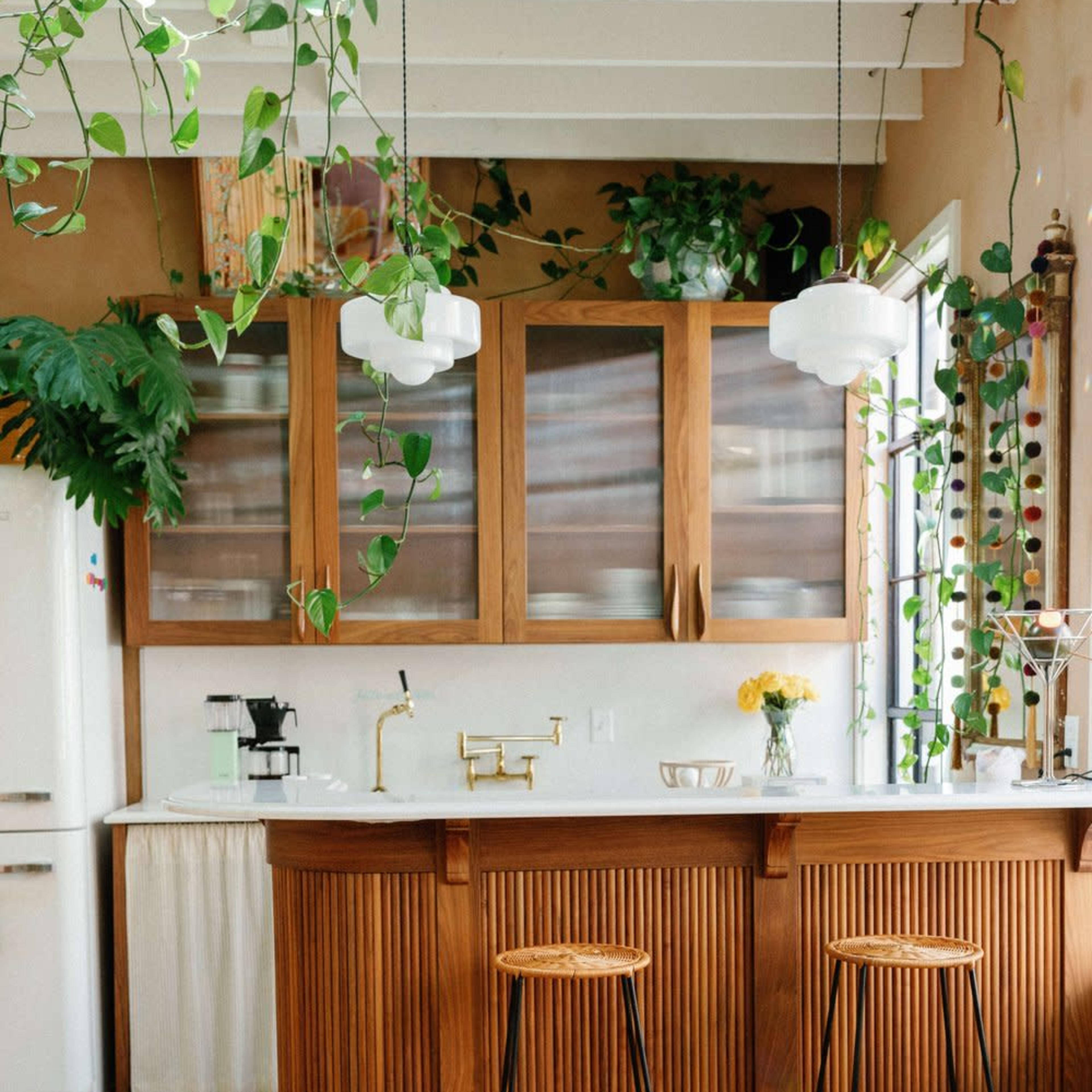 A modern kitchen features a wooden island with bar stools, surrounded by plants and natural light from large windows.