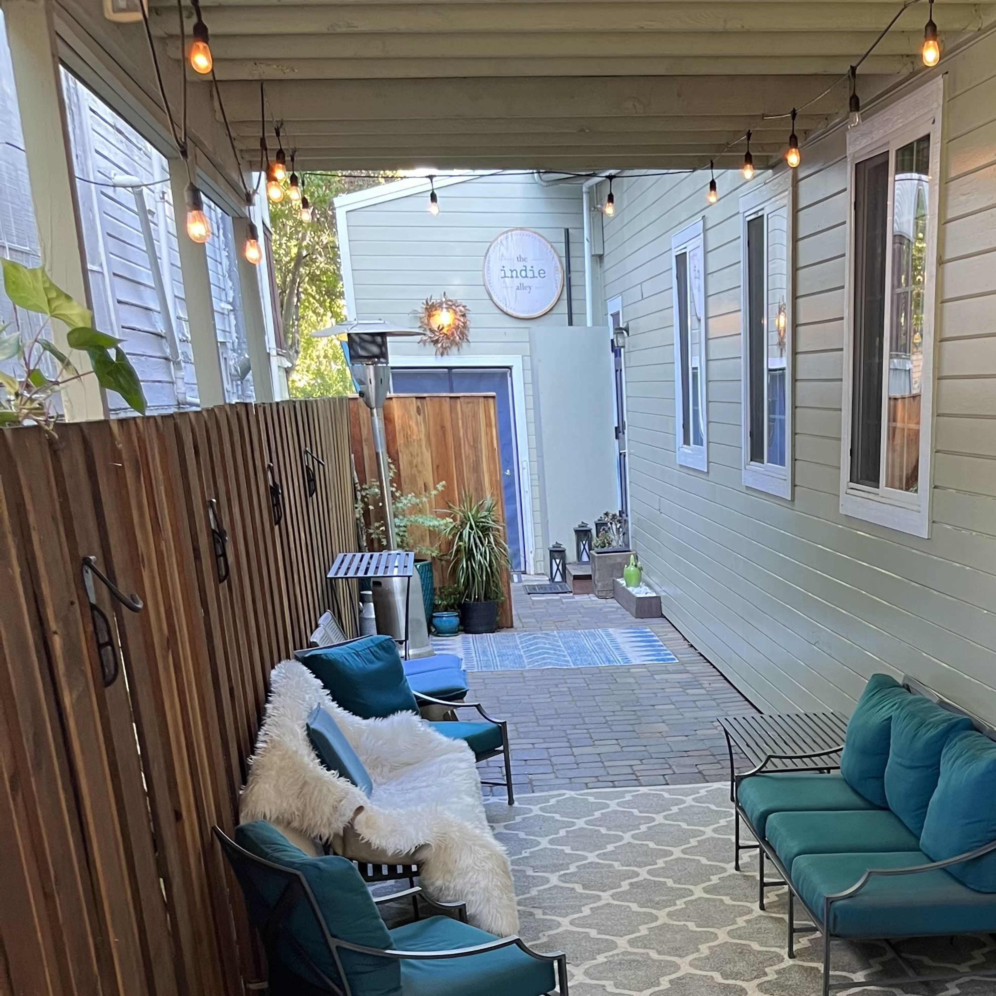 A narrow outdoor seating area lined with green walls, featuring two teal chairs, a rug, and string lights overhead.