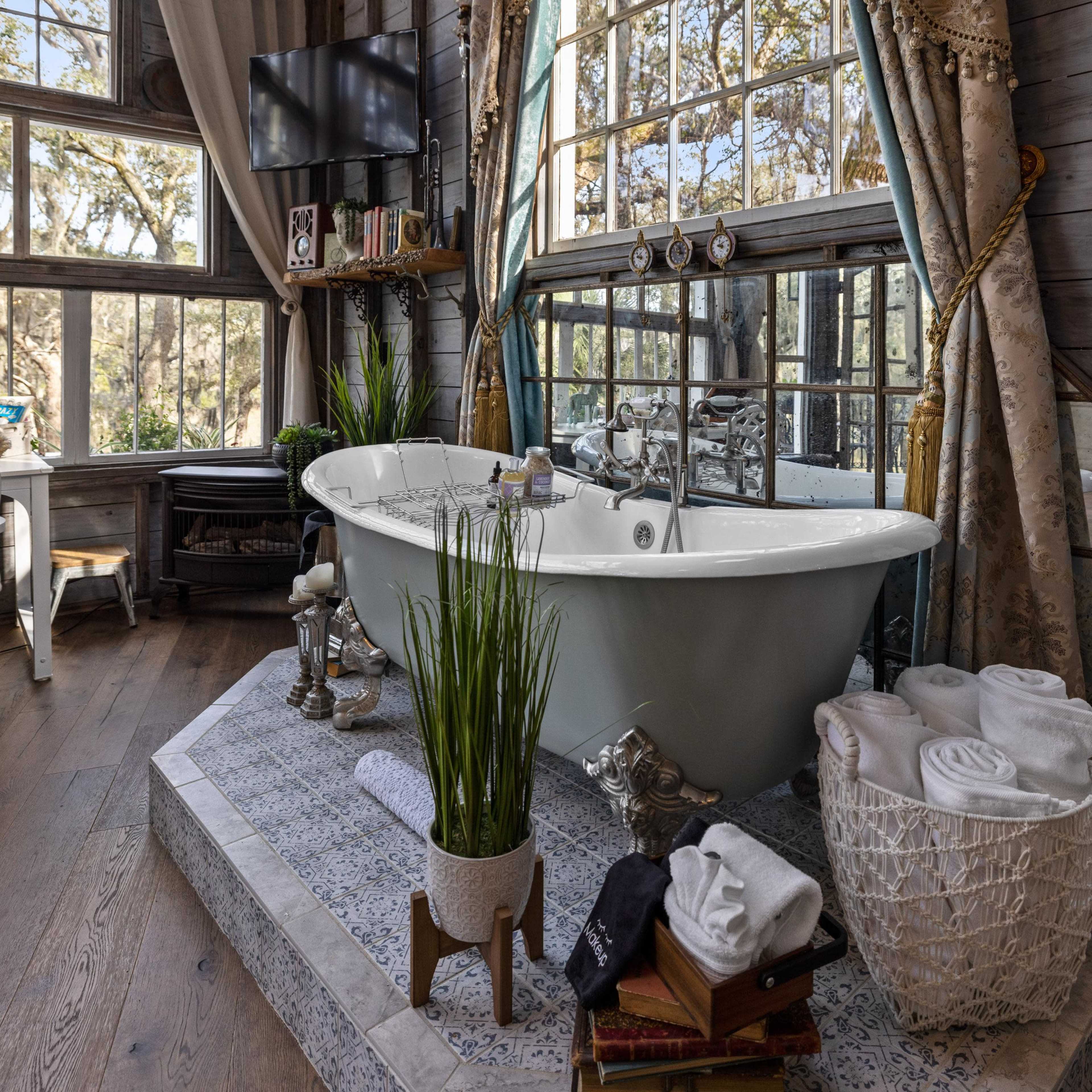 A spacious bathroom features a freestanding bathtub, large windows with natural light, and neatly arranged towels beside a plant and a small stack of books.