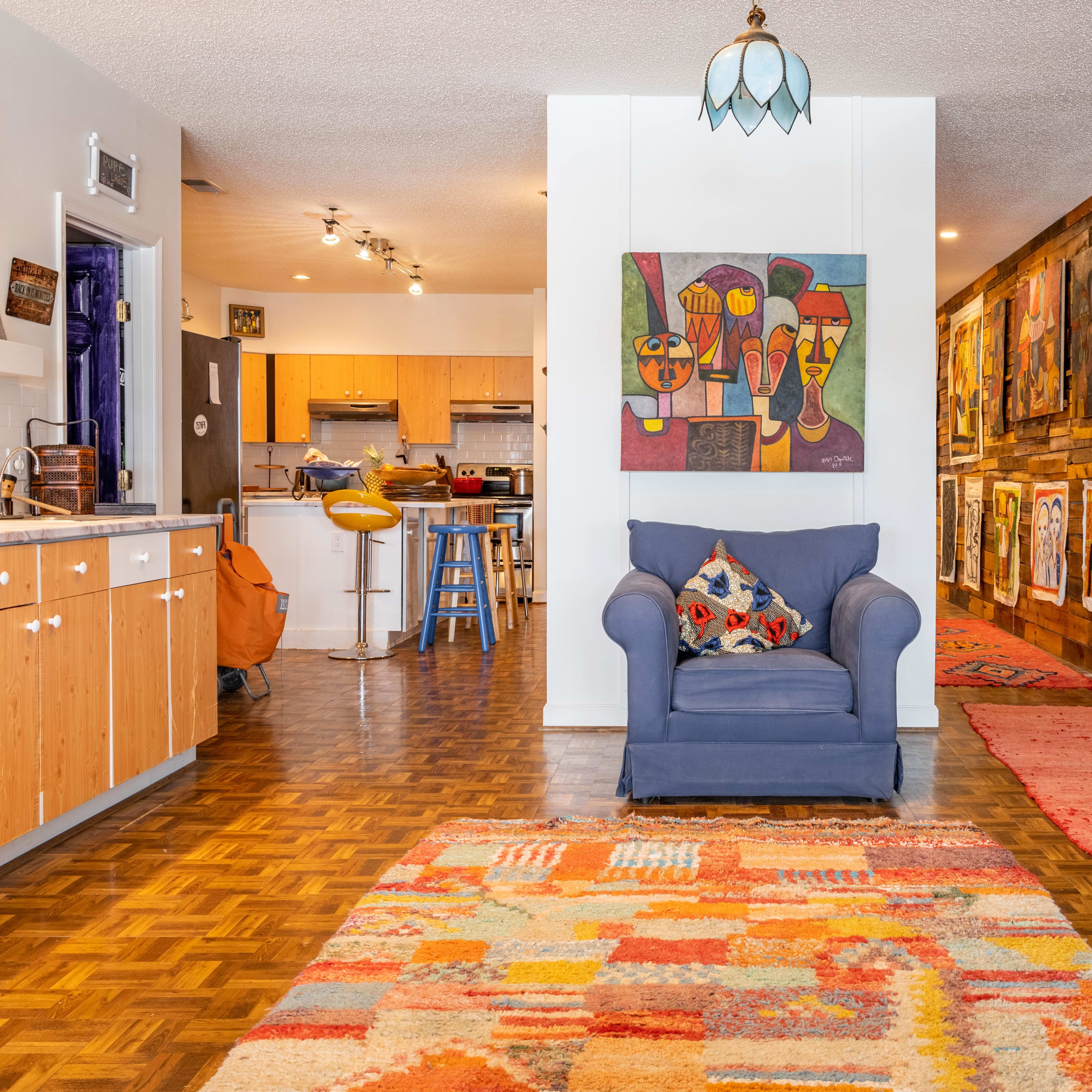 A spacious living area with a blue armchair, a wooden wall adorned with colorful artwork, and a patterned rug on the floor.