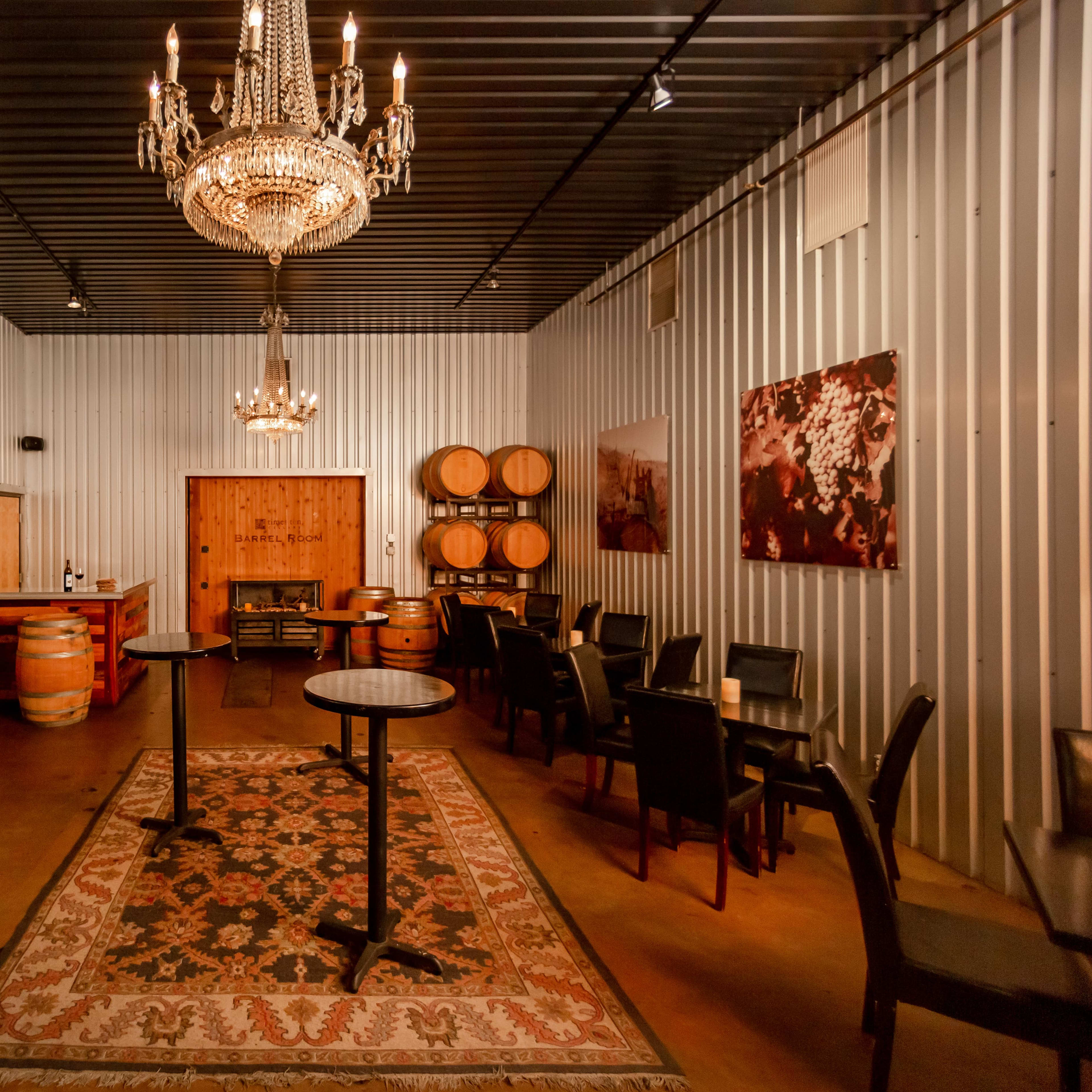 A dimly lit, metal-walled room furnished with tables, chairs, a chandelier, and wooden barrels.