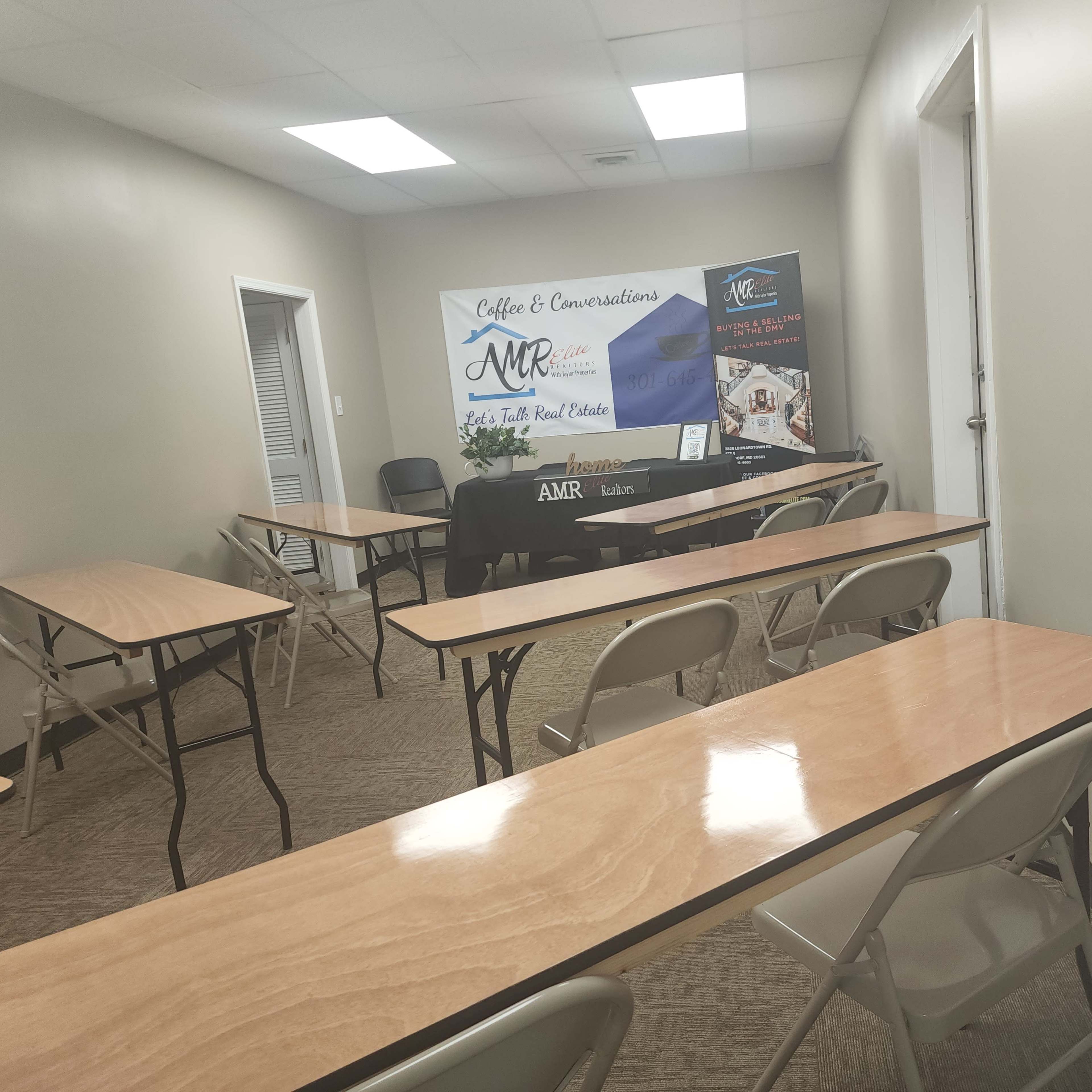 A sparsely furnished meeting room with several wooden tables and chairs arranged in a classroom style, featuring a promotional banner for "Coffee & Conversations" and "AMR" at the front.