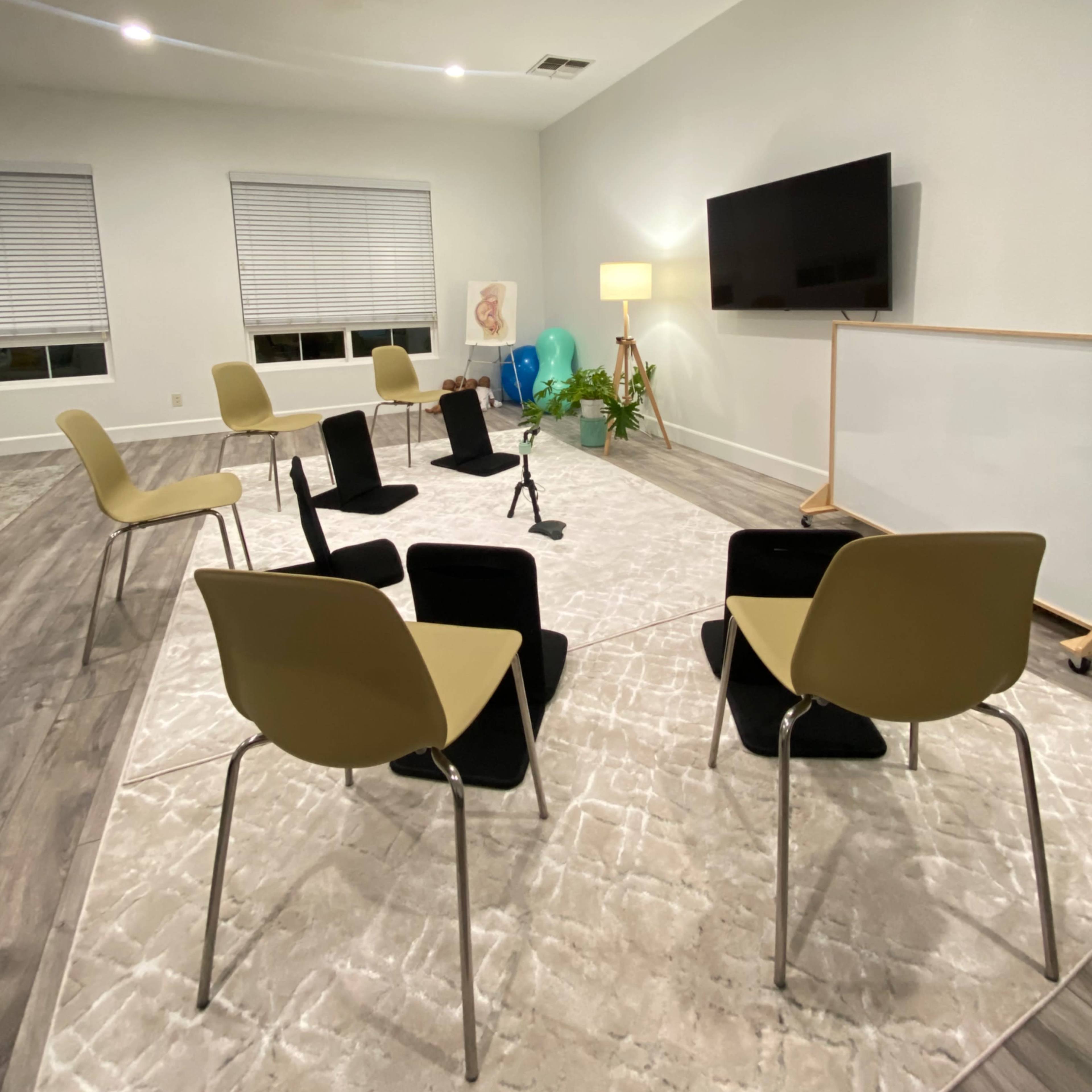 A room features six chairs arranged in a circle on a light-colored rug, with a television and whiteboard on opposing walls.