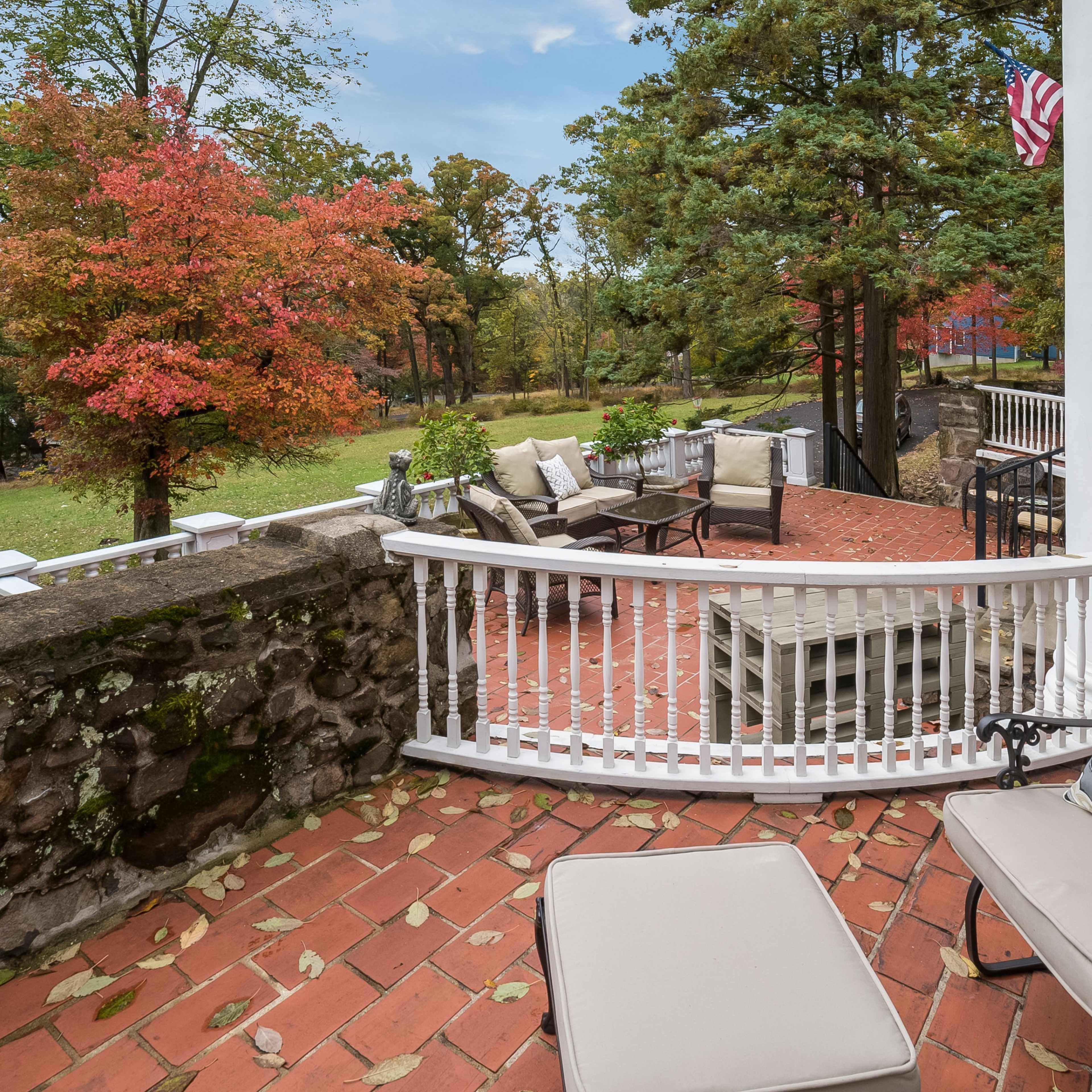 A brick patio with outdoor furniture overlooks a landscaped yard featuring autumn-colored trees and a stone wall.