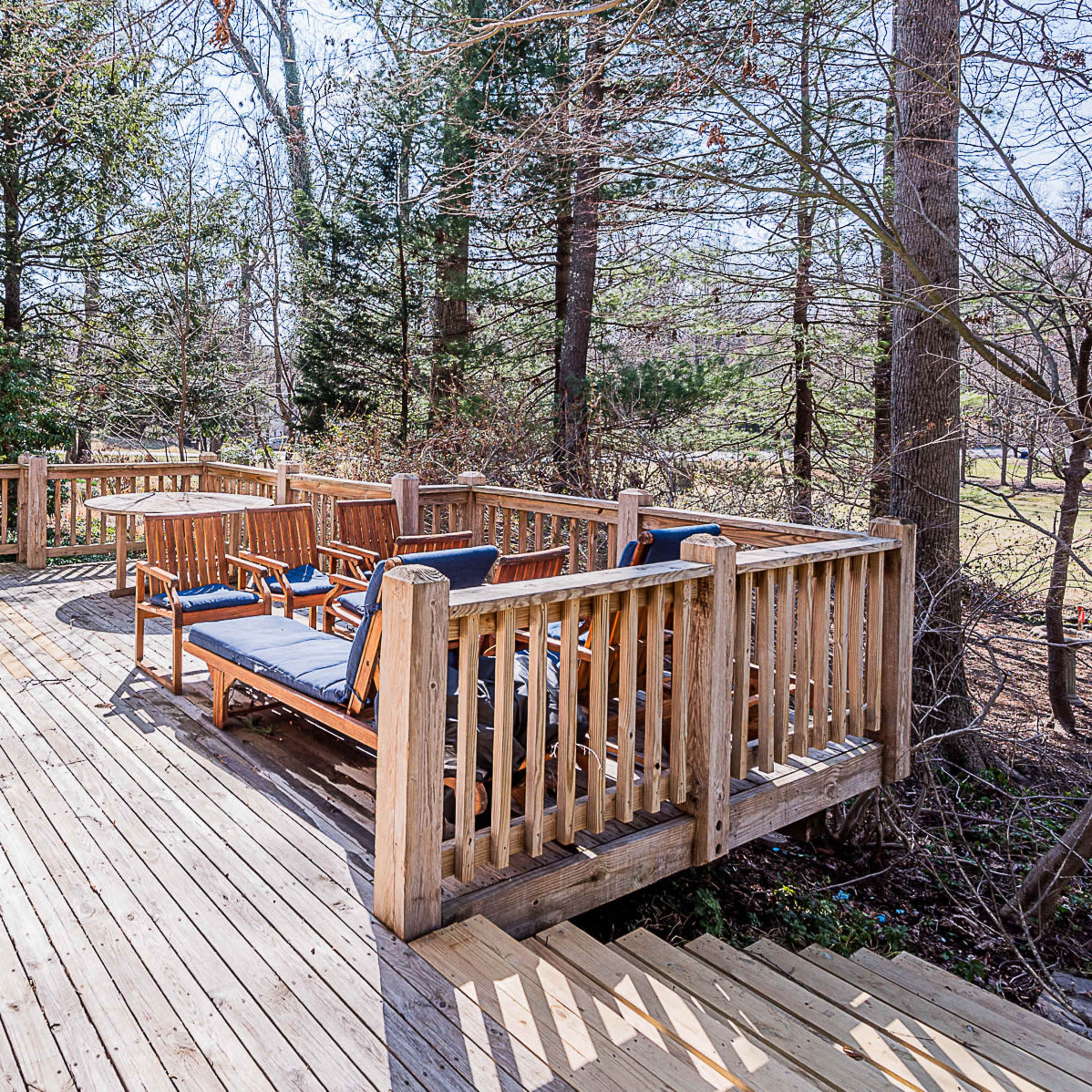 A wooden deck surrounded by trees, featuring several lounge chairs and a table.