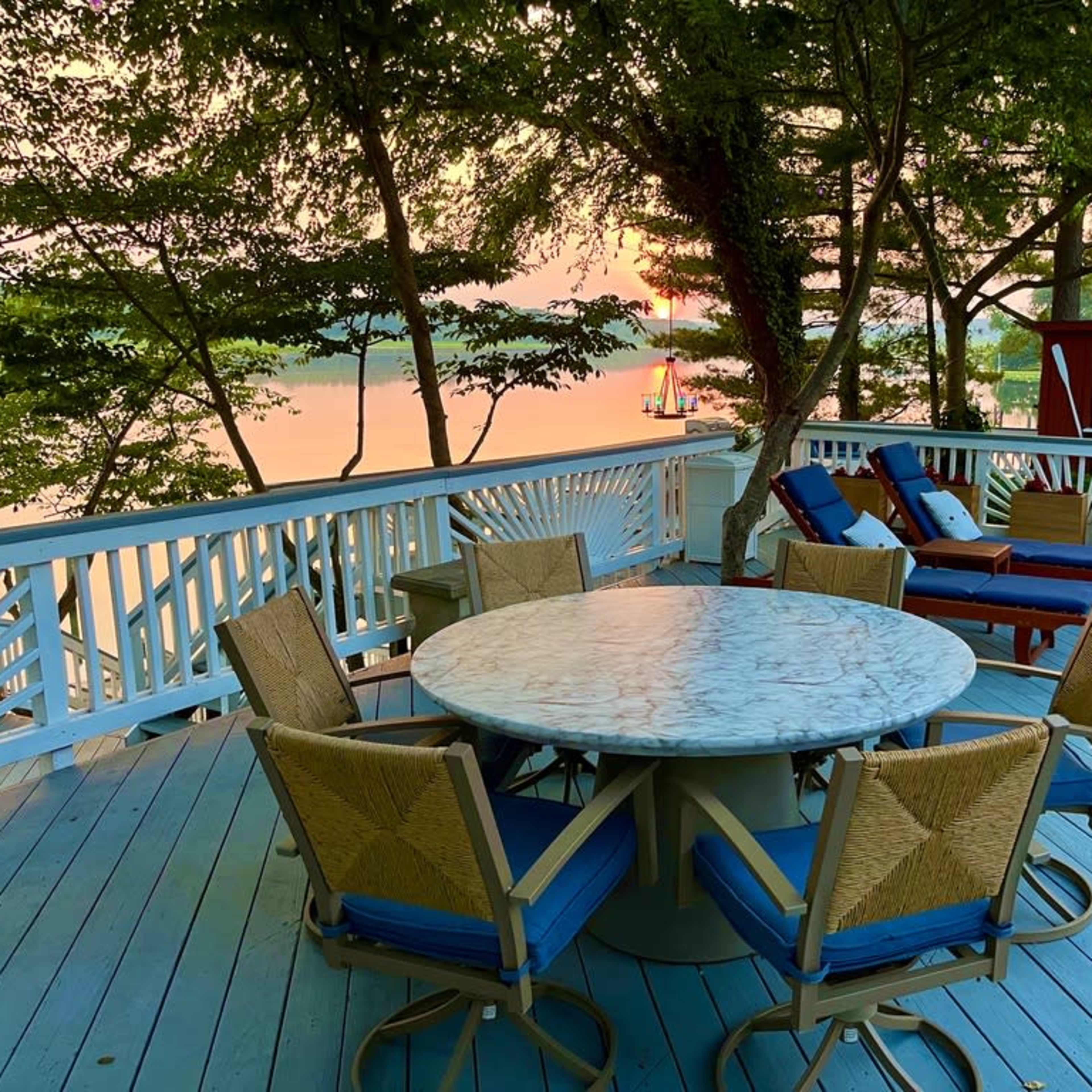 A circular marble table surrounded by eight chairs is set on a wooden deck overlooking a tranquil body of water at sunset.