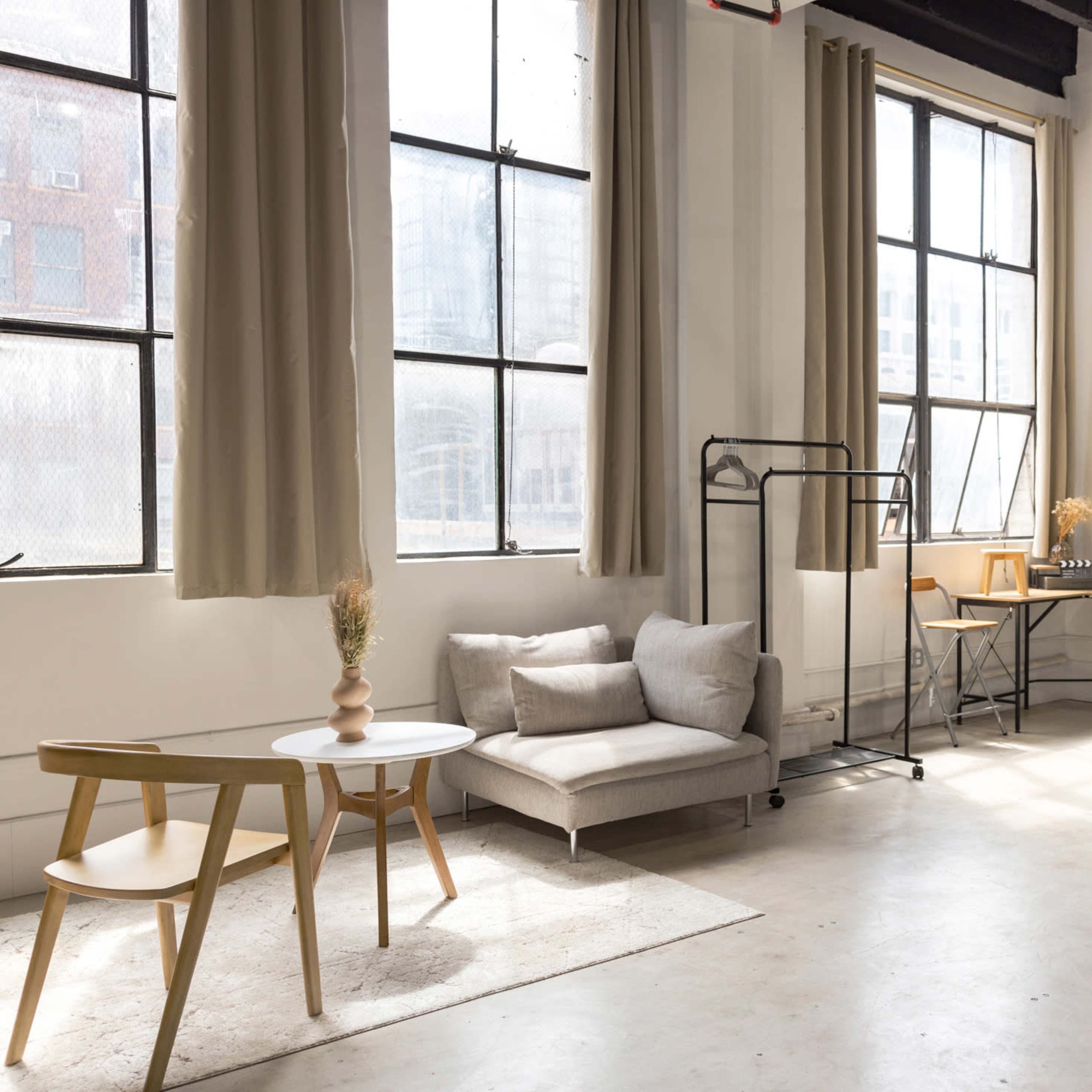 A spacious studio features large windows, minimalist furniture including a sofa and a table, and a workspace with a desk and chair.