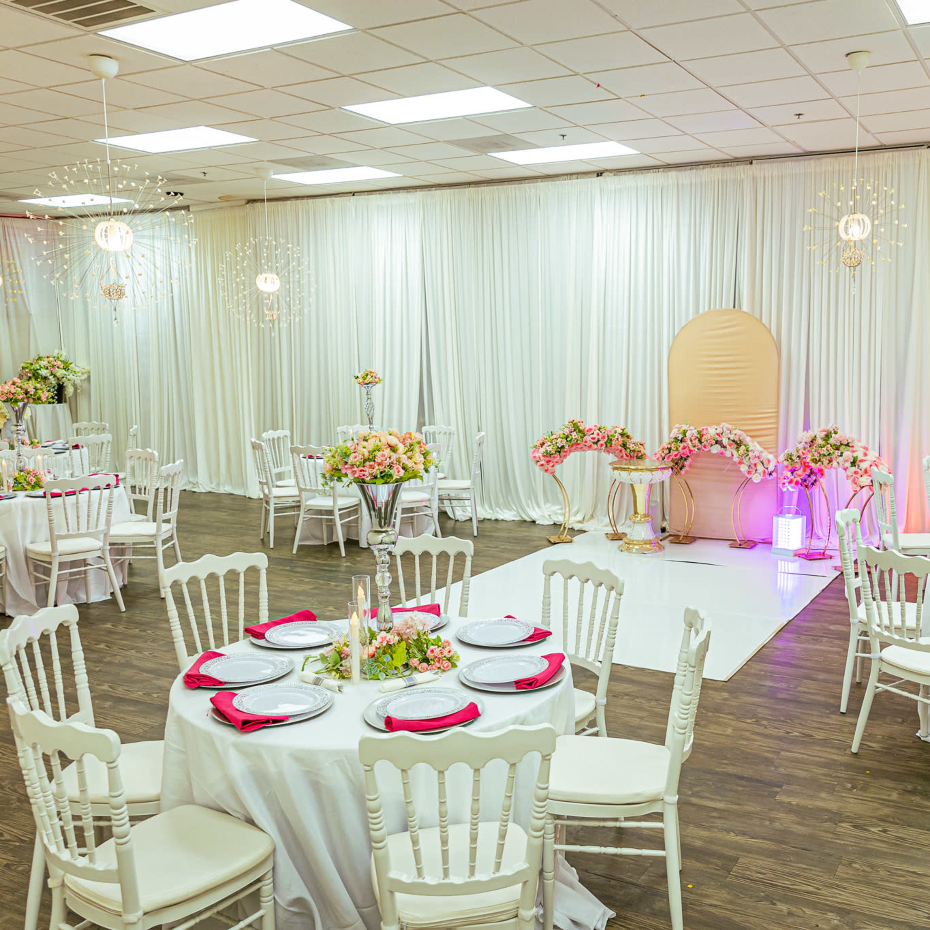 A banquet hall set up with round tables, each adorned with white tablecloths and pink accents, surrounded by floral arrangements and draped curtains.