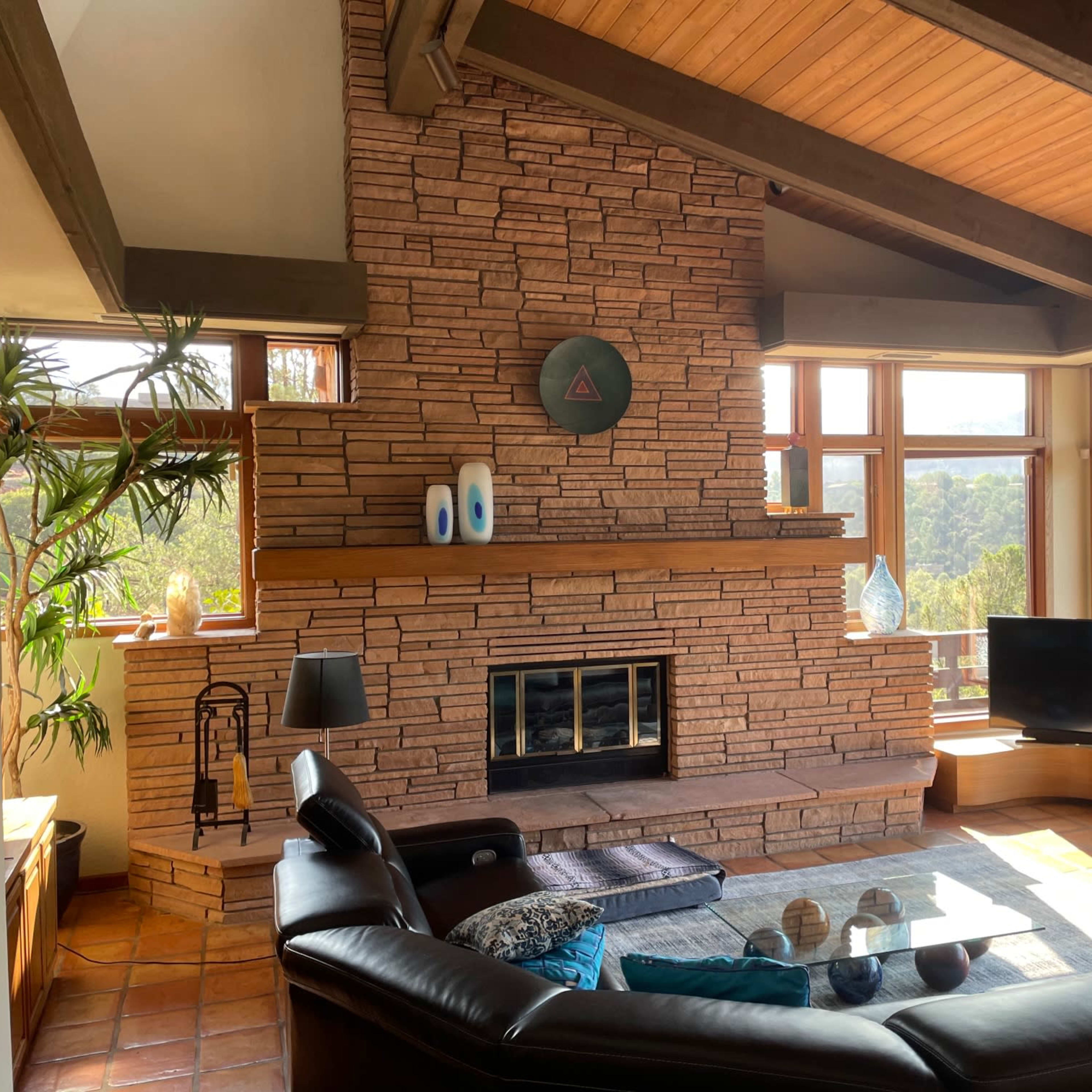 A living room with a stone fireplace, large windows offering a view of the outdoors, and a black leather sectional sofa.
