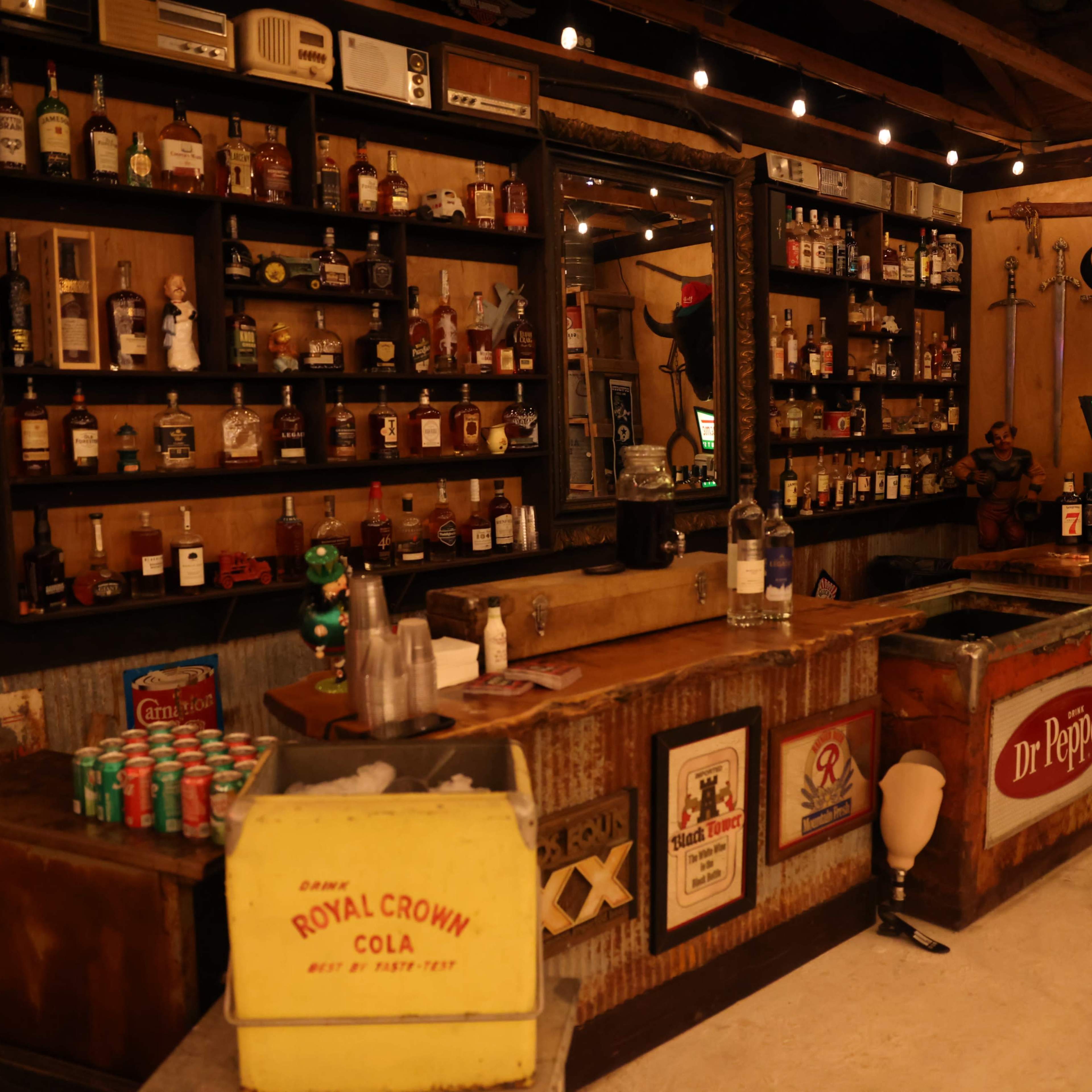 A rustic bar interior with shelves filled with various liquor bottles, vintage signs, and a wooden counter.